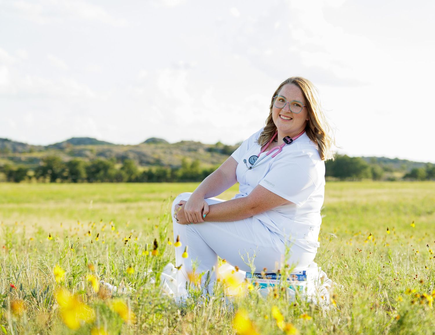 Smiling nursing student in white scrubs sitting in a sunny wildflower meadow with hills behind.