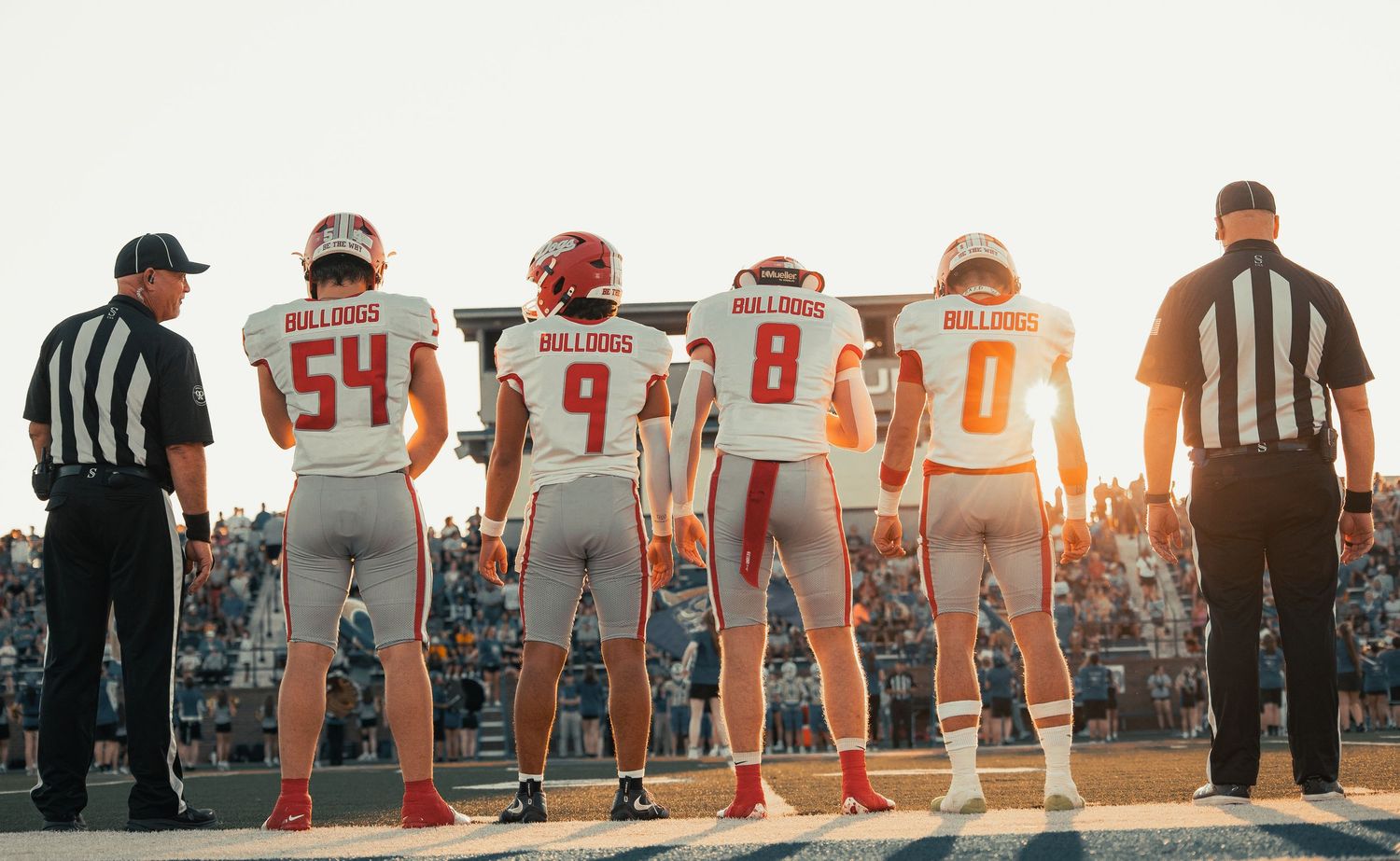 Four Bulldogs football players stand on the sideline in white uniforms, viewed from behind at sunset.