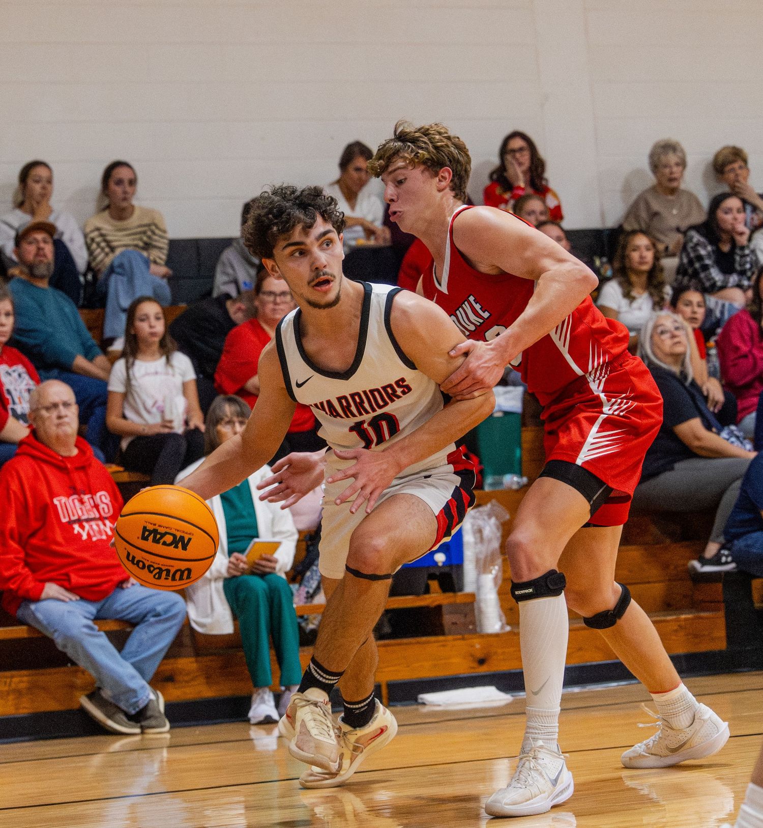 Warriors player #10 drives to the basket against a red-uniformed defender in a packed high school basketball gym.