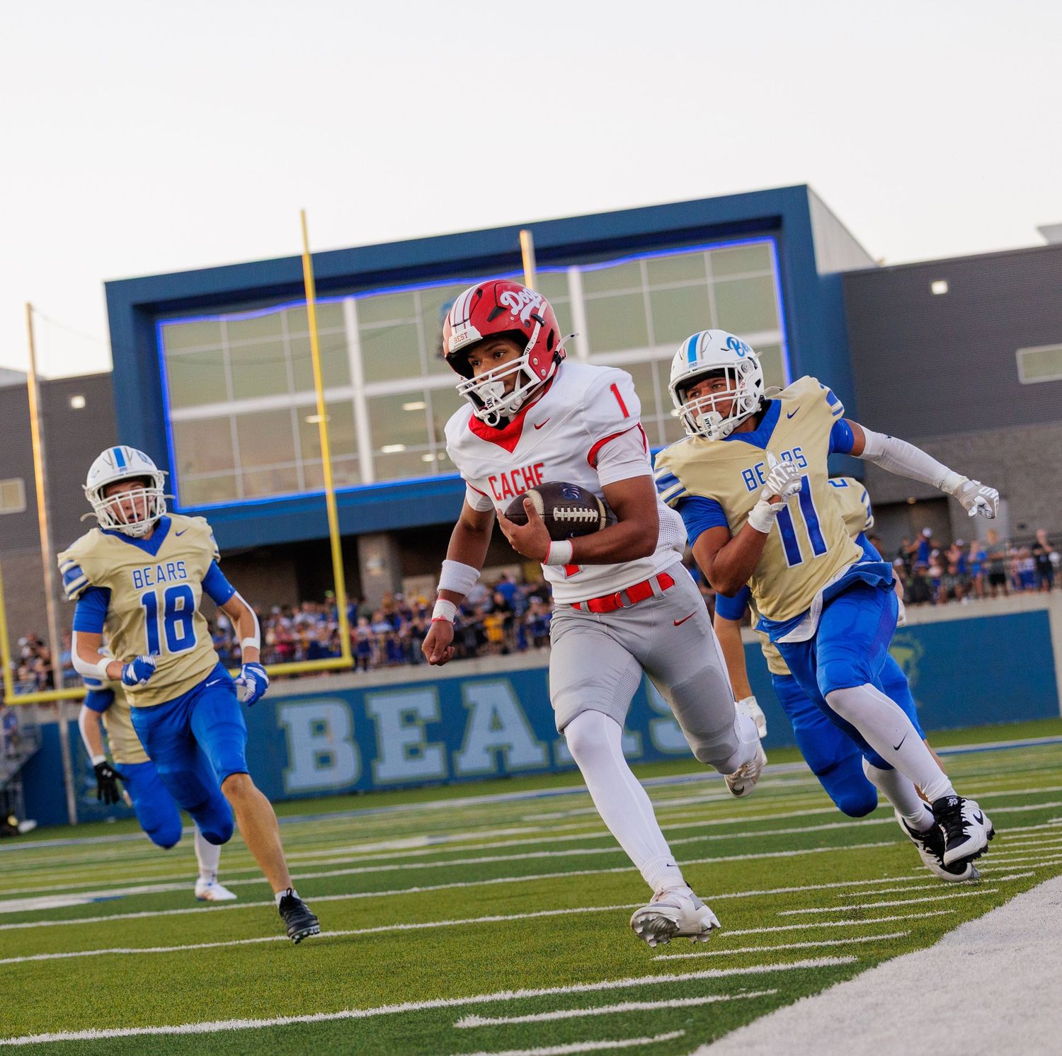 A football player in red uniform runs toward the end zone while blue-jersey defenders give chase during a night game.