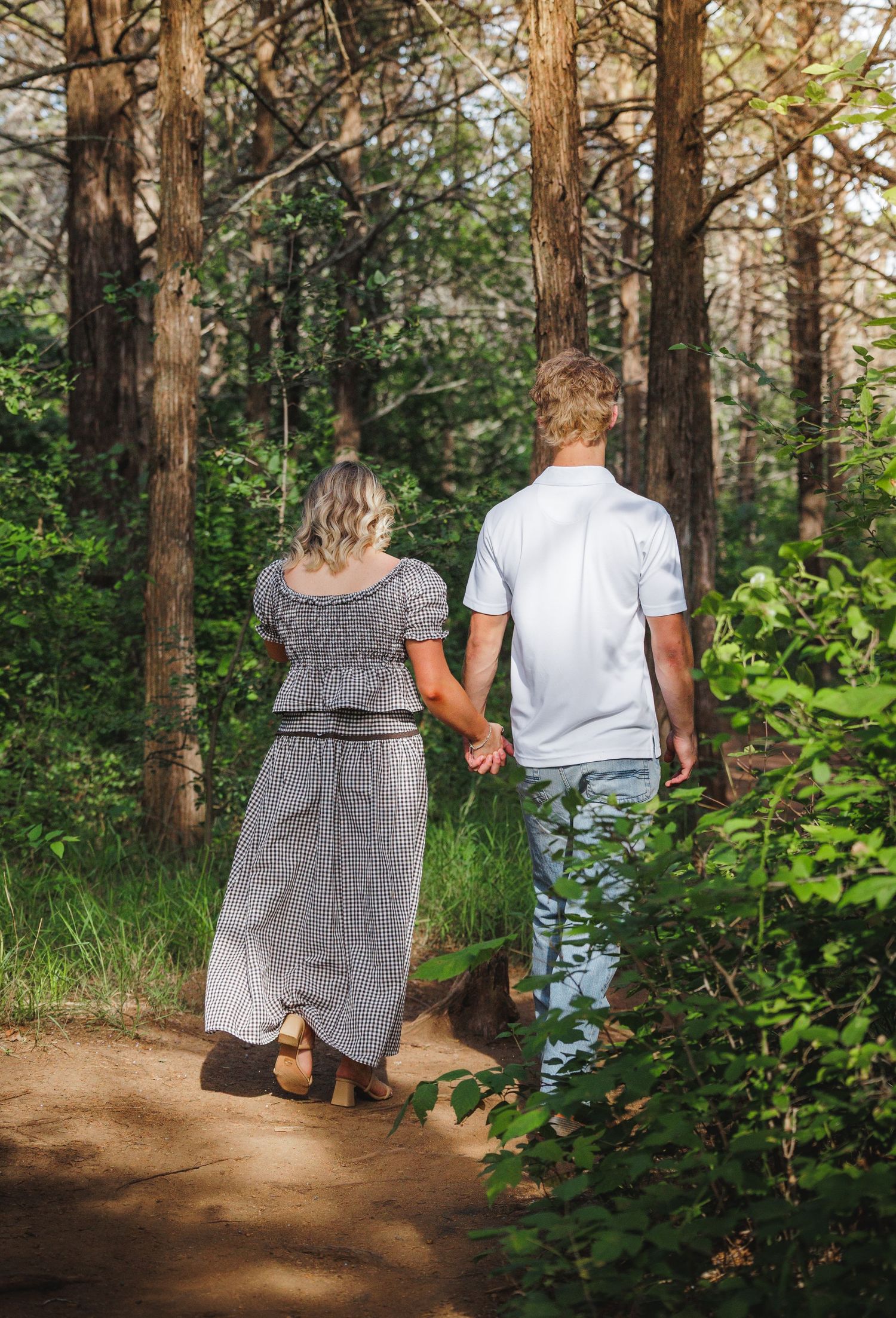 Couple holding hands walking on a forest path surrounded by tall trees and lush green foliage.