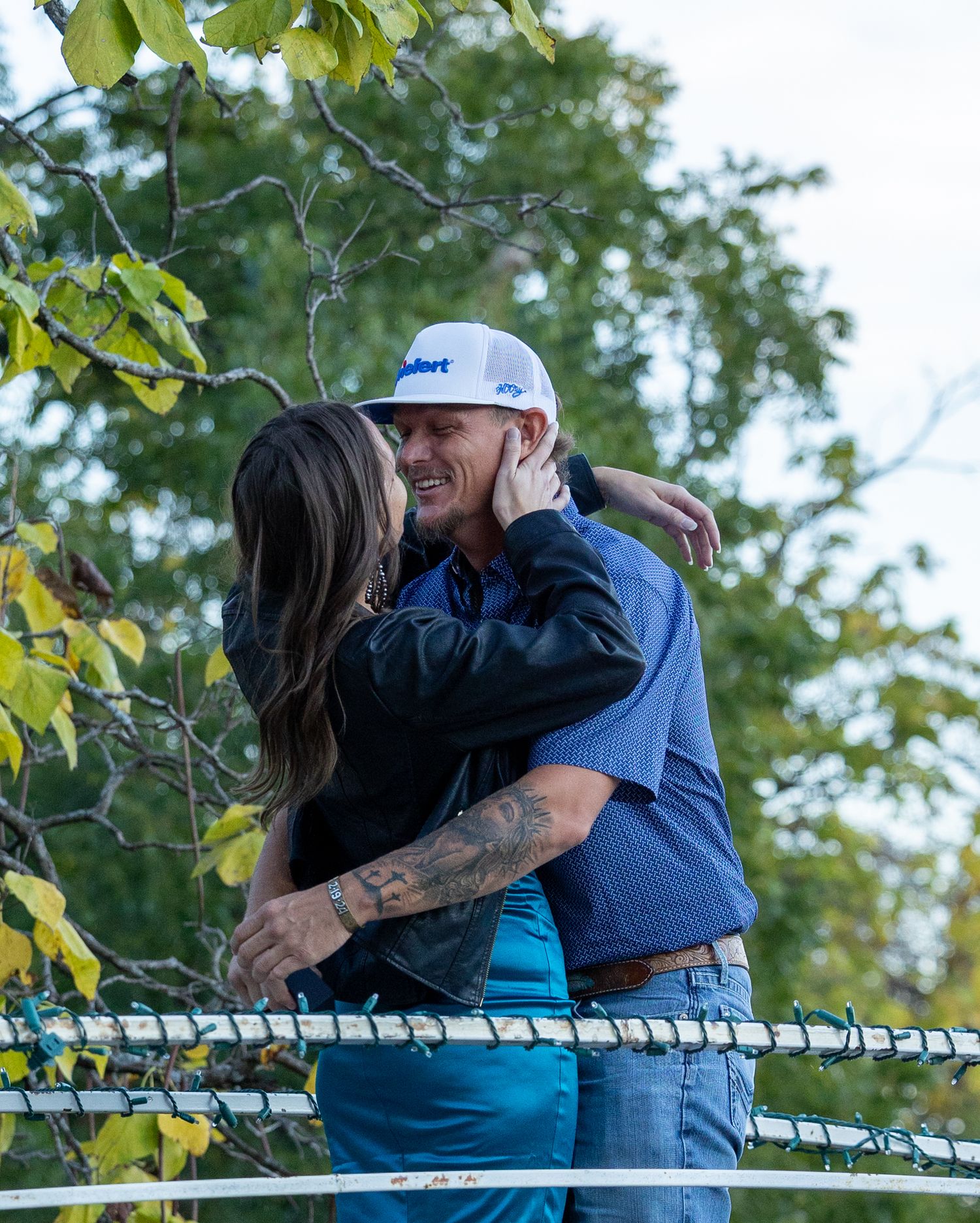 Couple shares a romantic kiss while embracing outdoors near a fence surrounded by green trees.