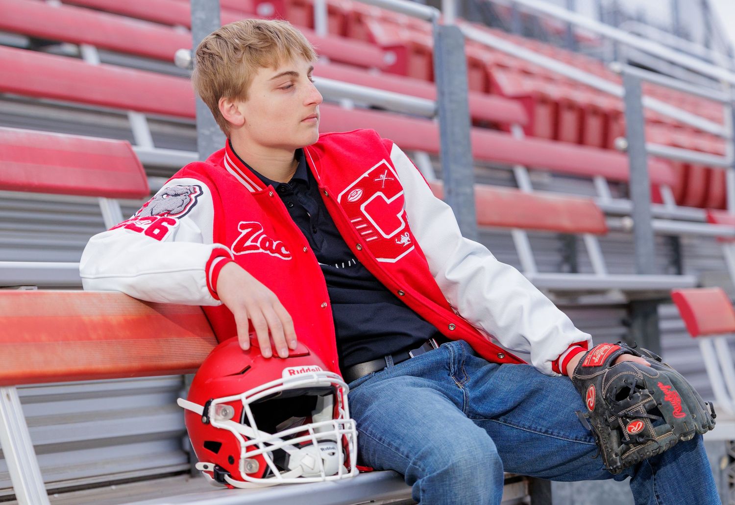 A teen in a red letterman jacket rests in bleachers with a football helmet and baseball glove.