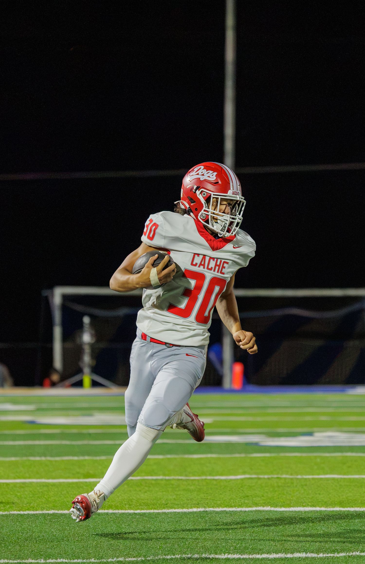 Cache High School football player #30 in red and white uniform runs with the ball during a night game.