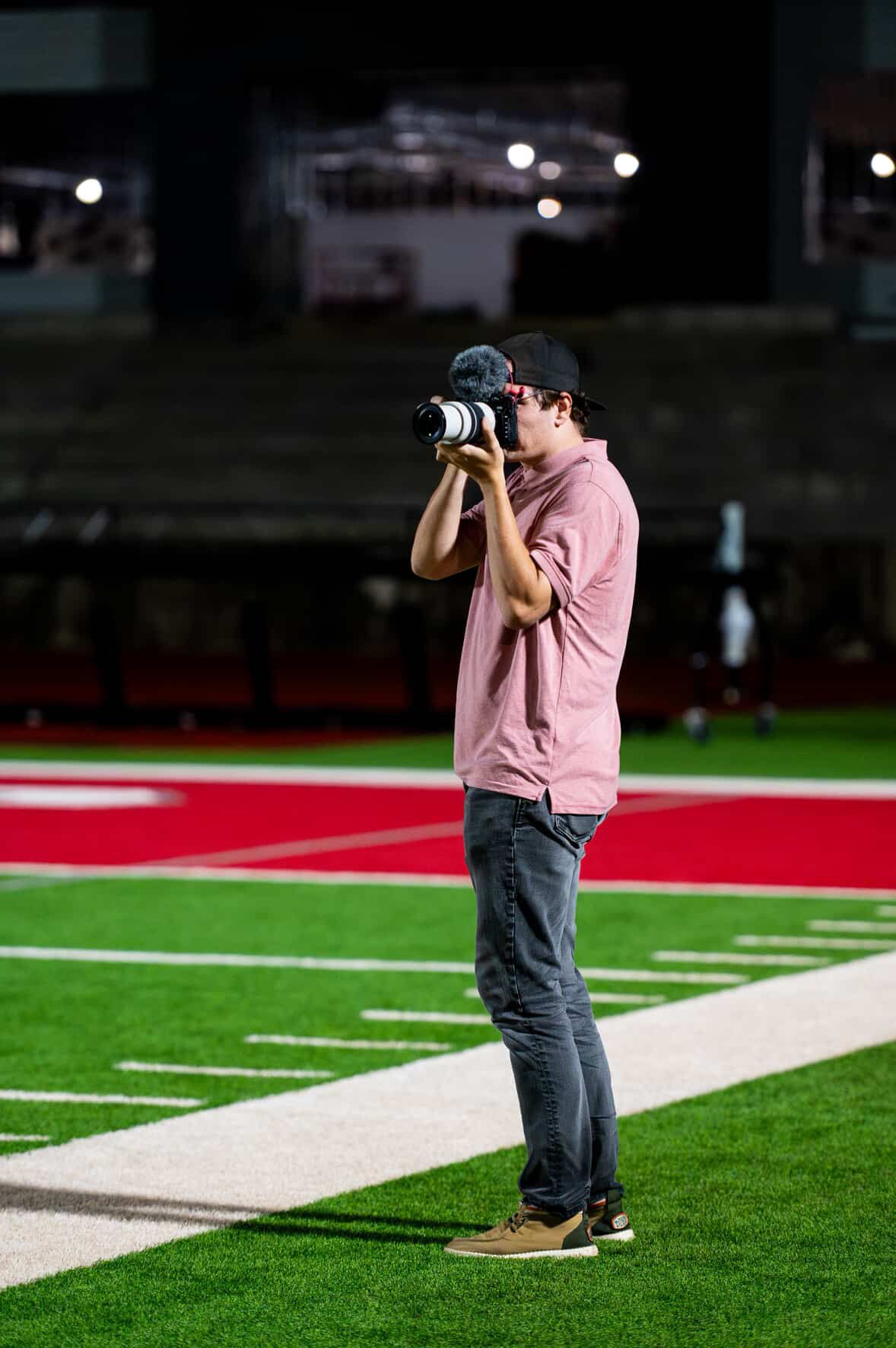 Male photographer in pink shirt using a camera on a red football field at night.