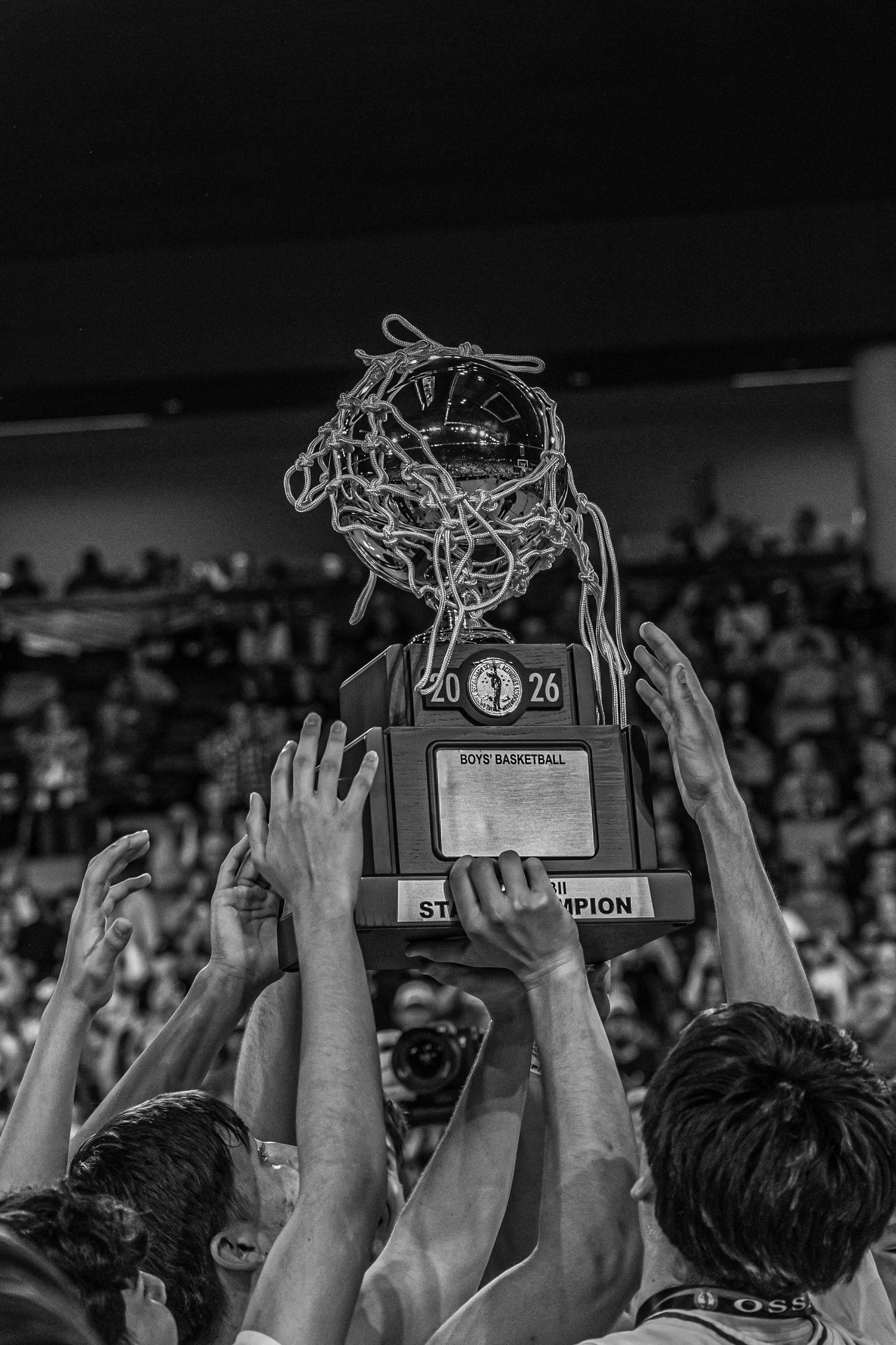 Hands raised holding a Boys Basketball State Champion trophy with a net draped over it in black and white.
