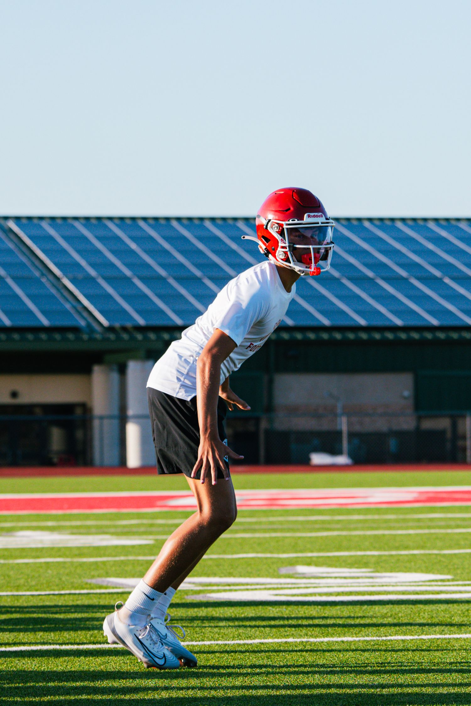 Young football player in red helmet and white jersey crouches in a ready stance during outdoor field practice.