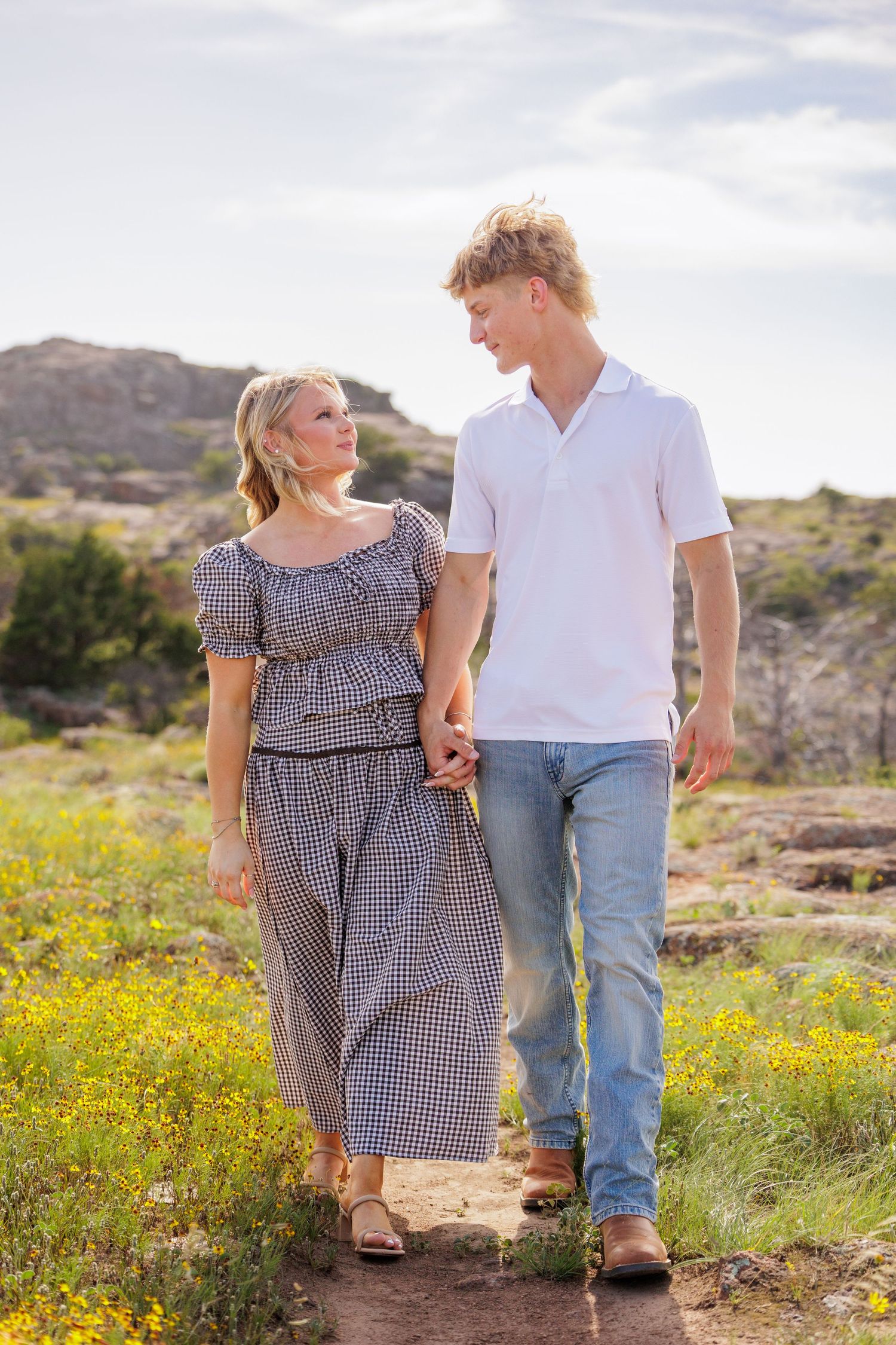 A young couple holds hands while walking through a scenic wildflower meadow with rocky hills under a bright sky.