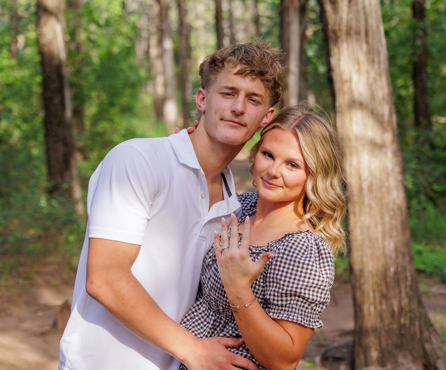 Young couple embracing outdoors in a forest setting, woman showing engagement ring, wearing casual summer attire.