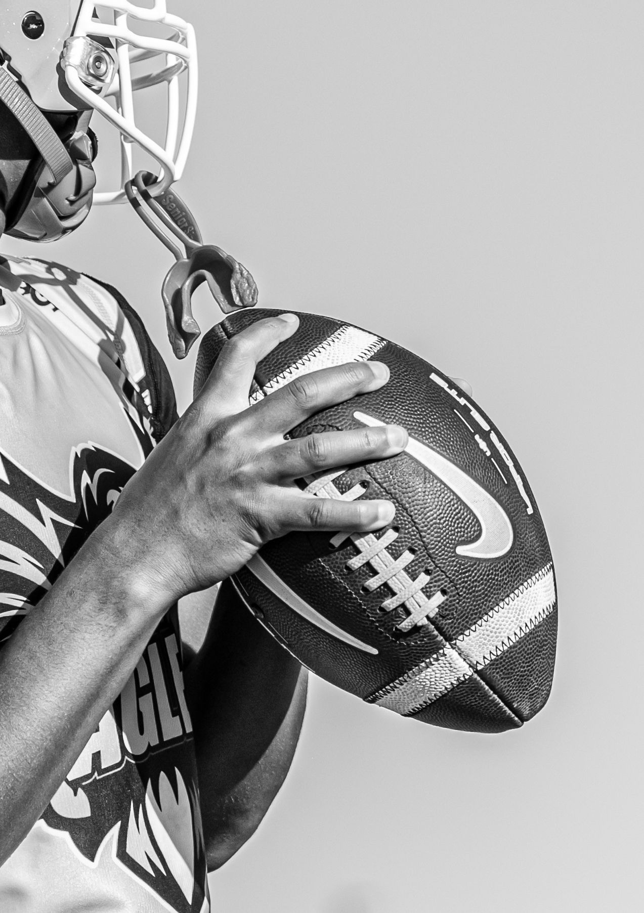 Close-up black and white photo of a football player gripping a Nike football while wearing a helmet.