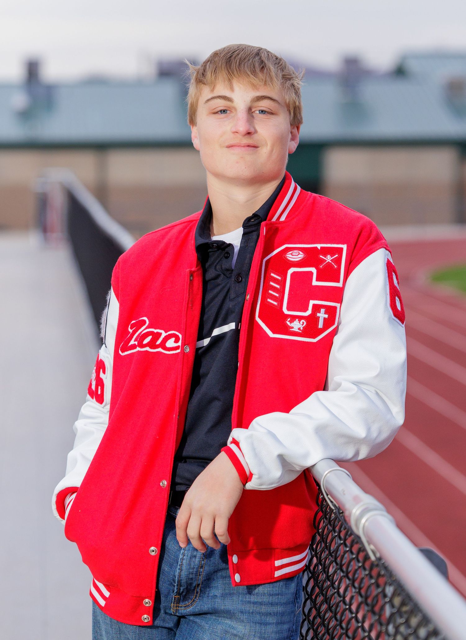 A young athlete poses in a red and white letterman jacket leaning on a railing at a stadium.