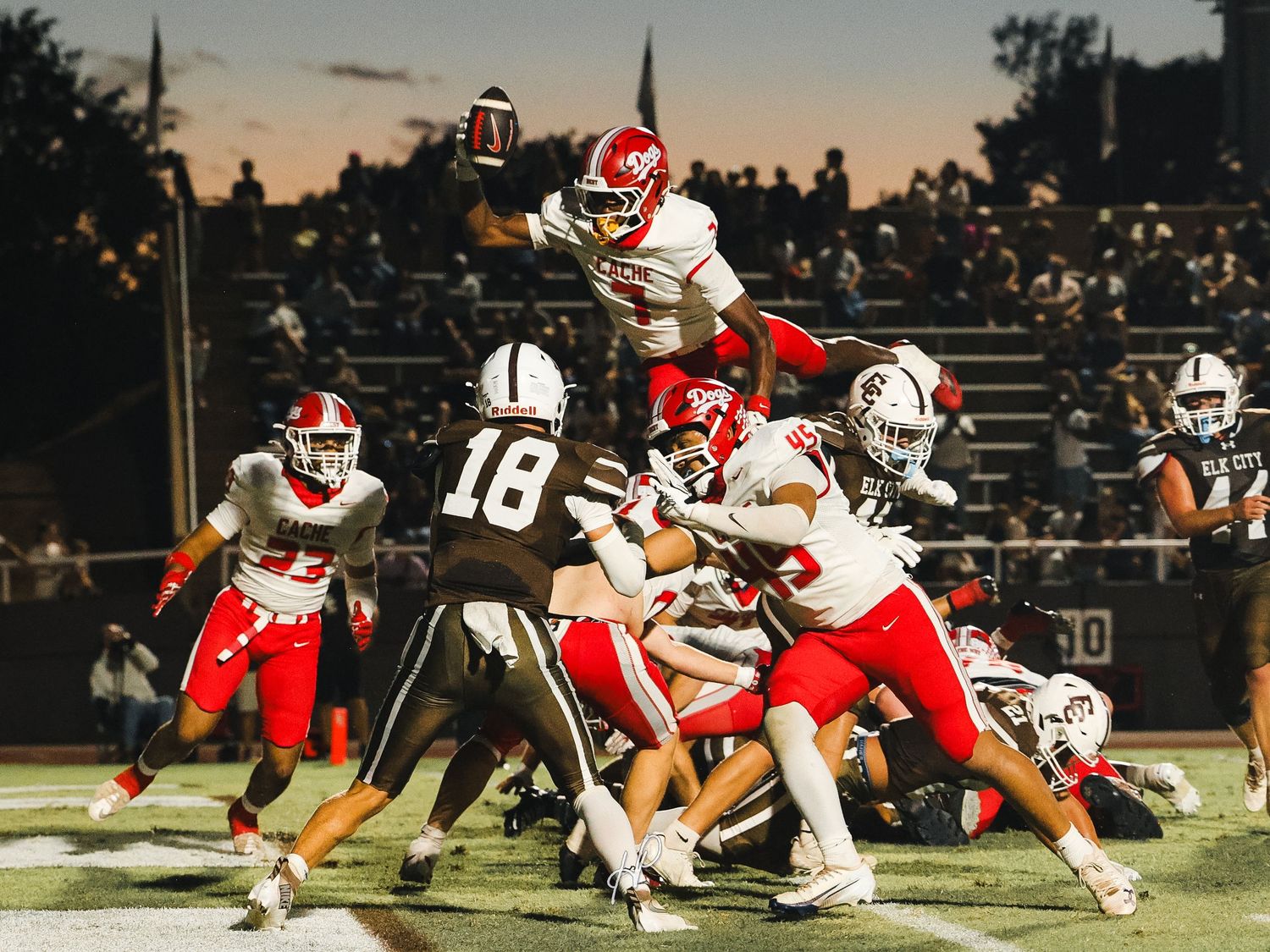 Football player leaps over defensive line during intense high school night game with red and white uniforms.
