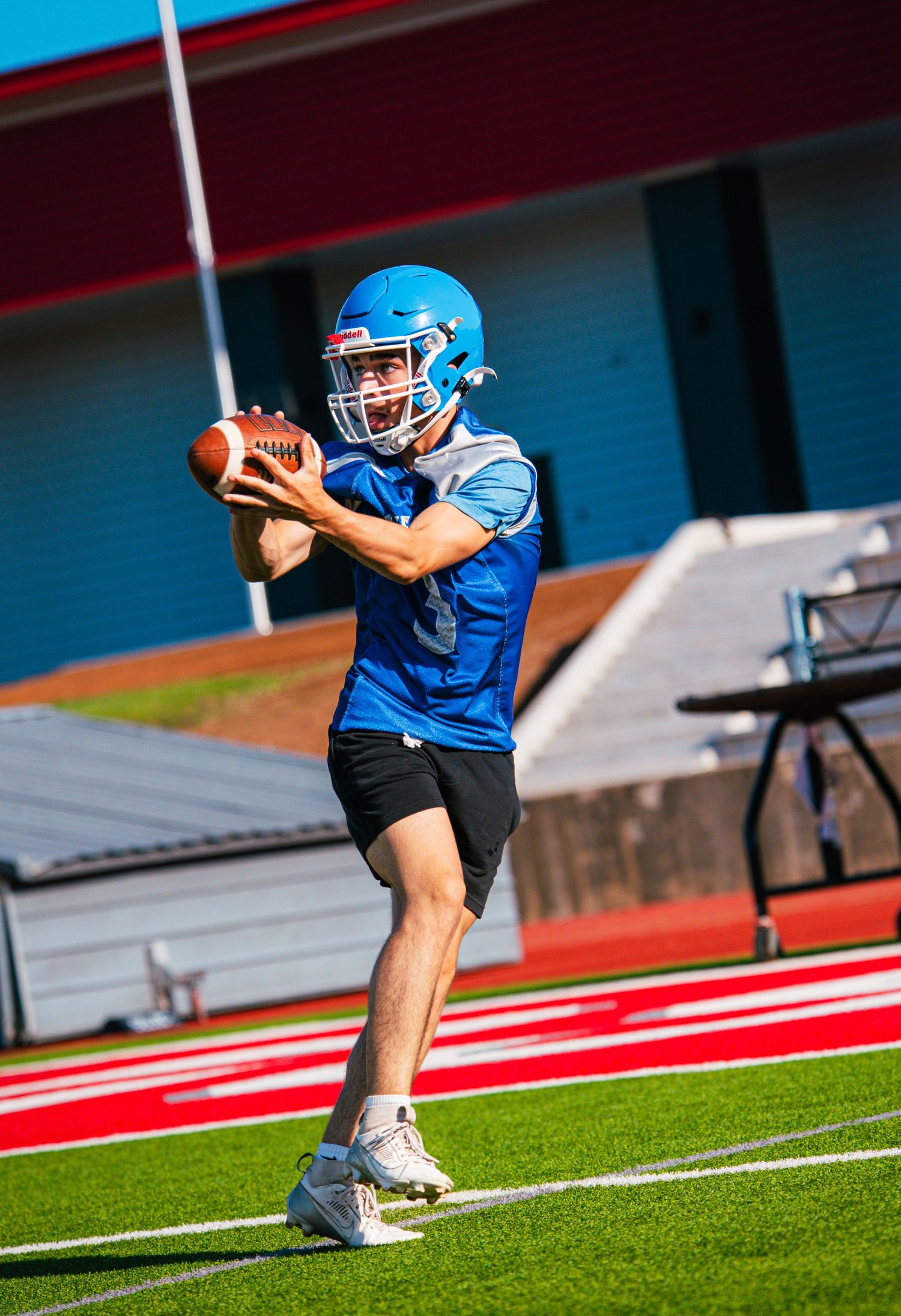 Young football player in blue helmet and jersey prepares to throw a football during stadium practice.