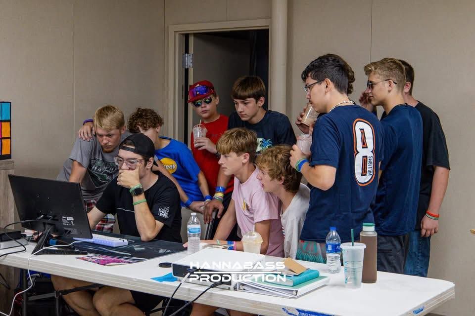 Group of teenage boys gathered around a laptop computer at a table in a classroom setting.