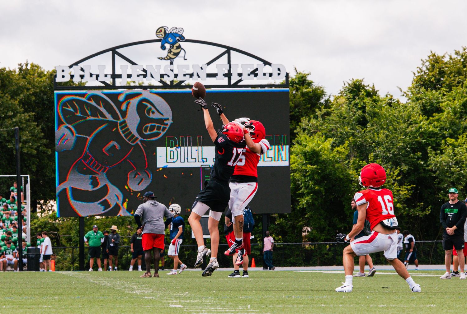 Football players jump to catch a pass during an outdoor practice game on a green field with a scoreboard in the background.
