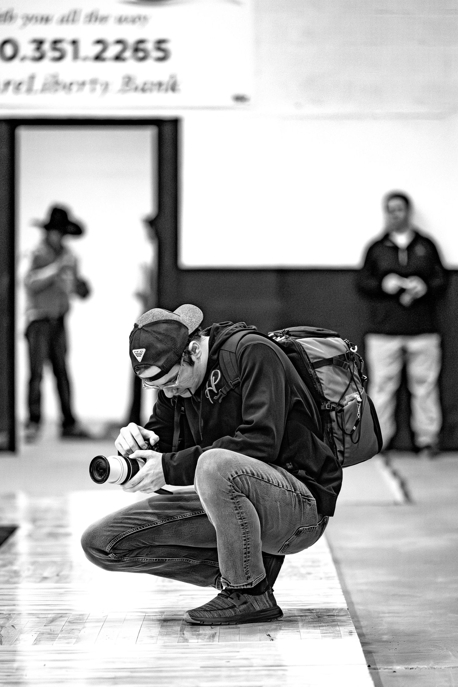 Black and white photo of a photographer crouching to take a low-angle shot inside a sports gymnasium.