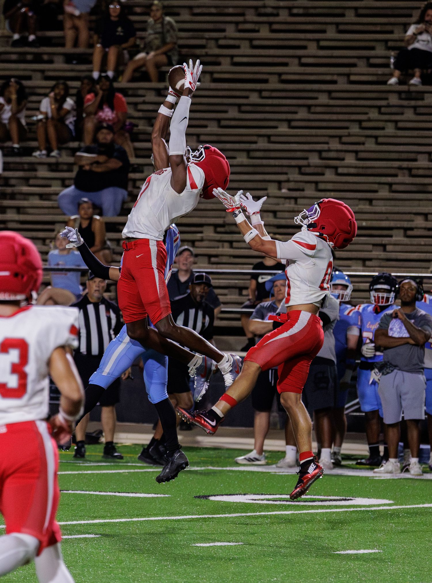 Two football players in red uniforms leap to catch a pass during a nighttime high school football game.
