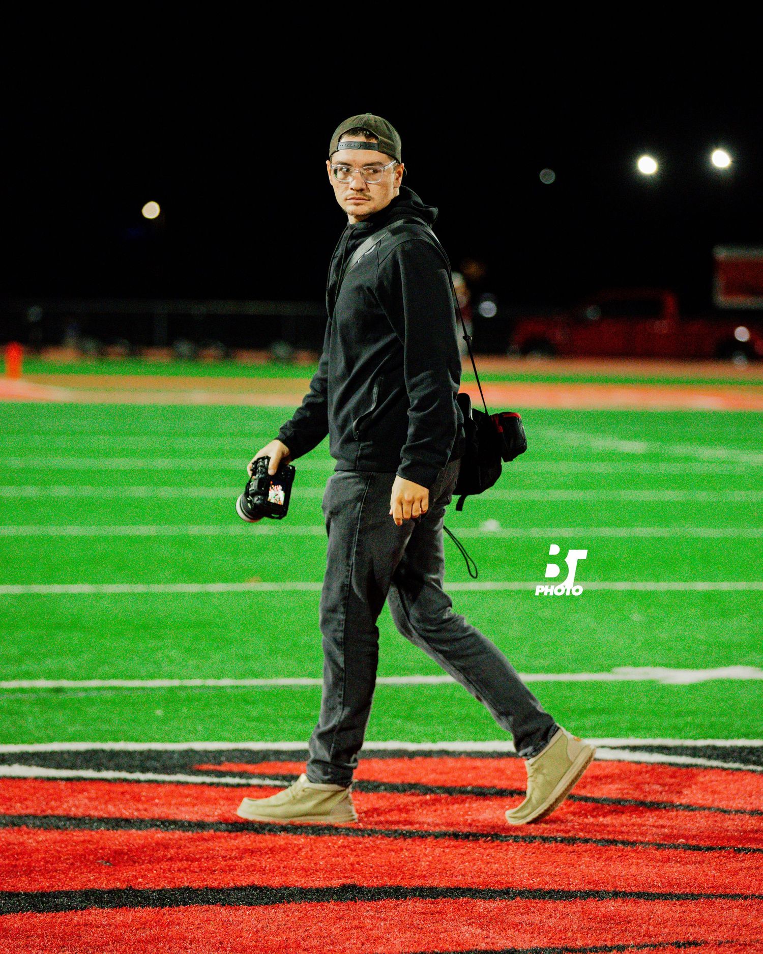 A photographer walks across a red and green football field at night, wearing all black with tan shoes and carrying camera equipment.