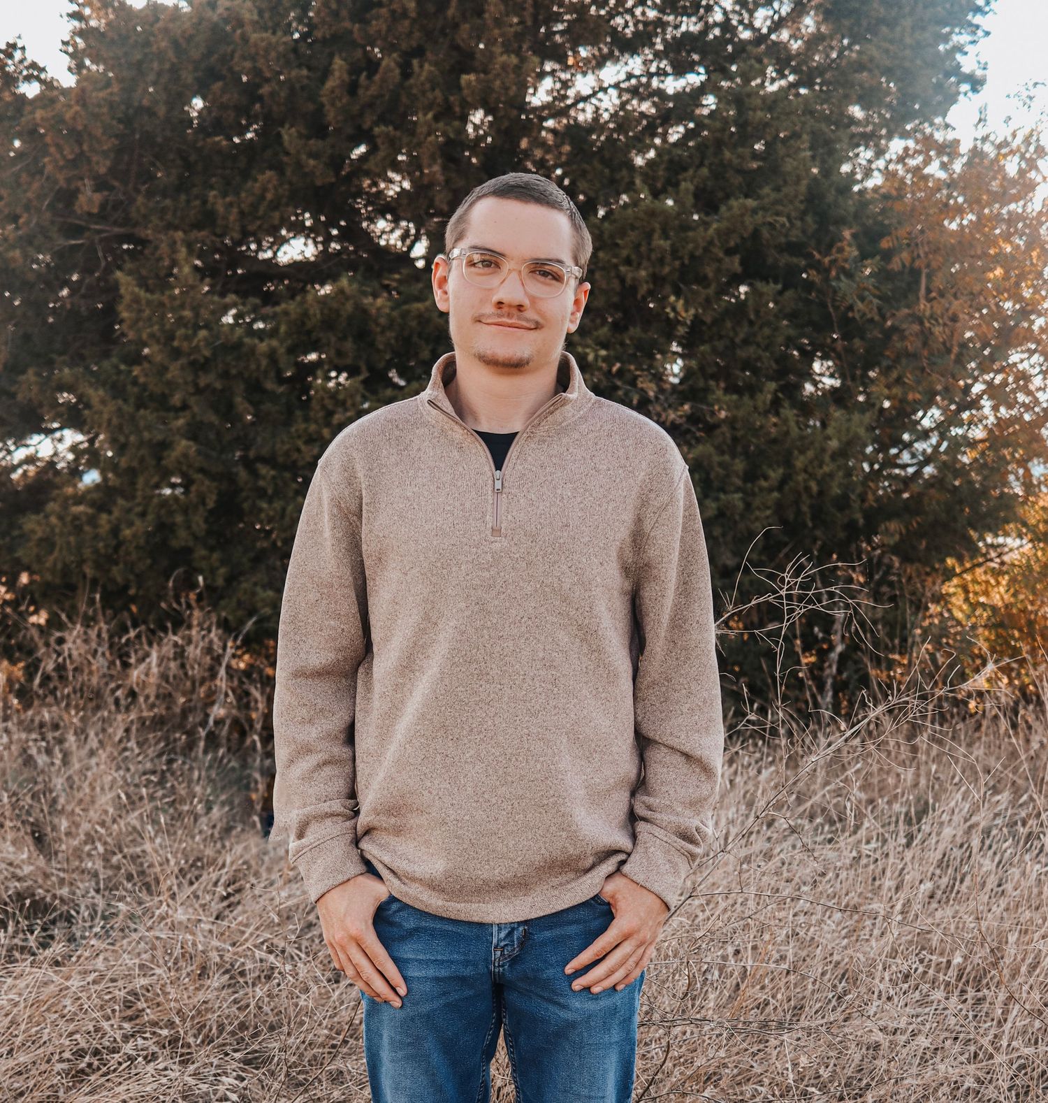 Young man in beige quarter-zip pullover stands outdoors in a field with trees at golden hour.