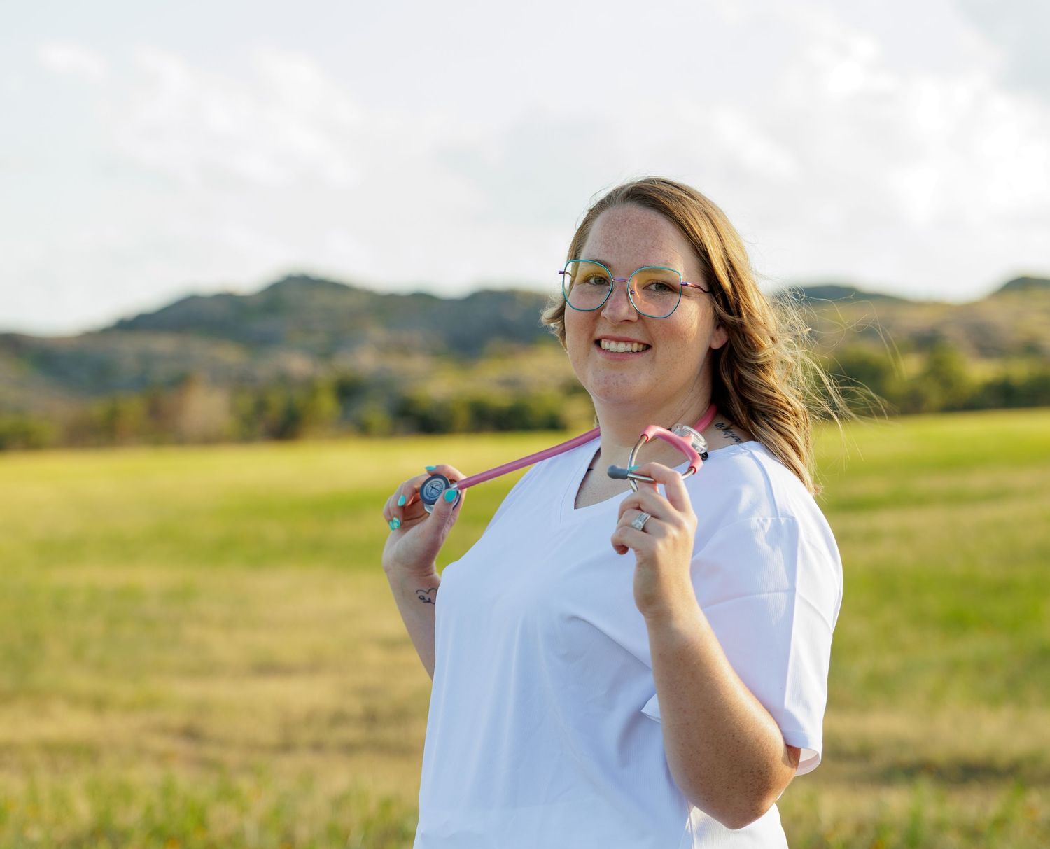 Smiling young woman in white scrubs holding a stethoscope in an outdoor field setting.