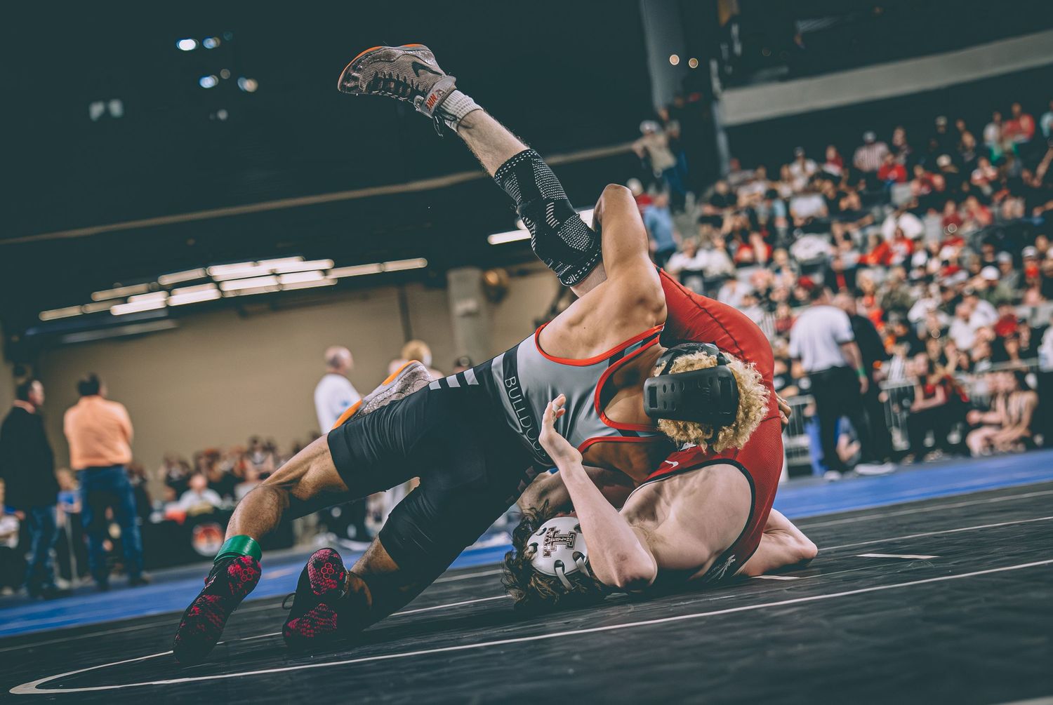 Two wrestlers compete on a mat during an indoor wrestling tournament with spectators in the background.