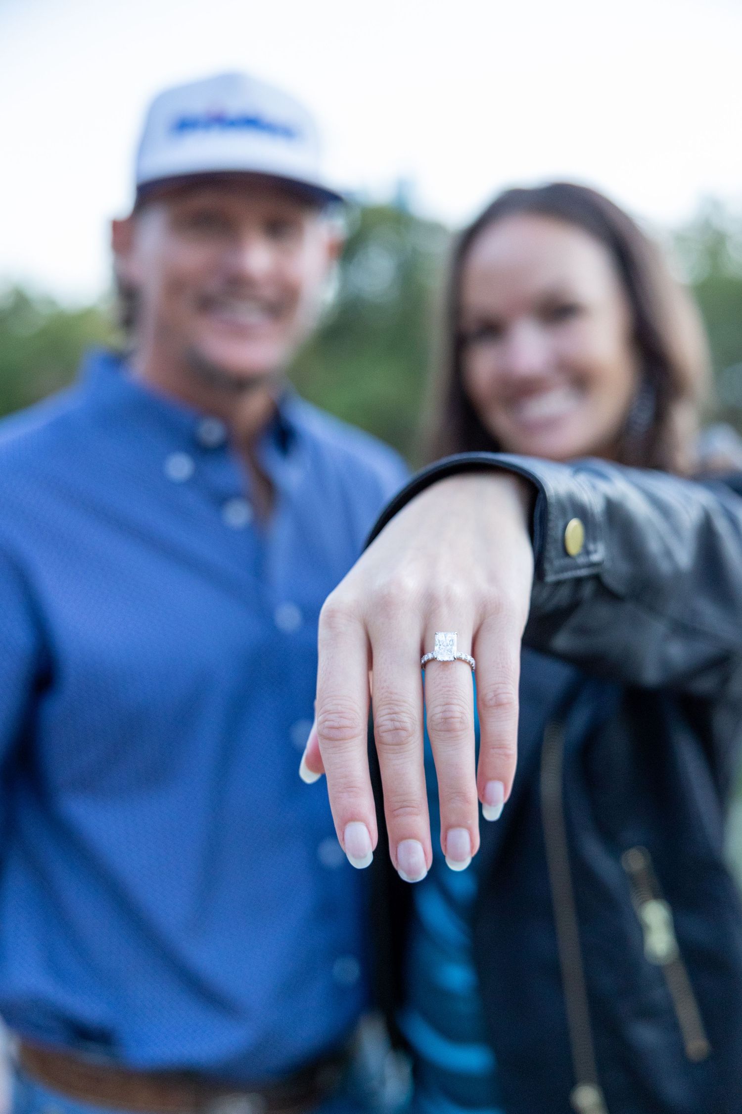 Newly engaged couple smiles while showing off a diamond engagement ring outdoors.
