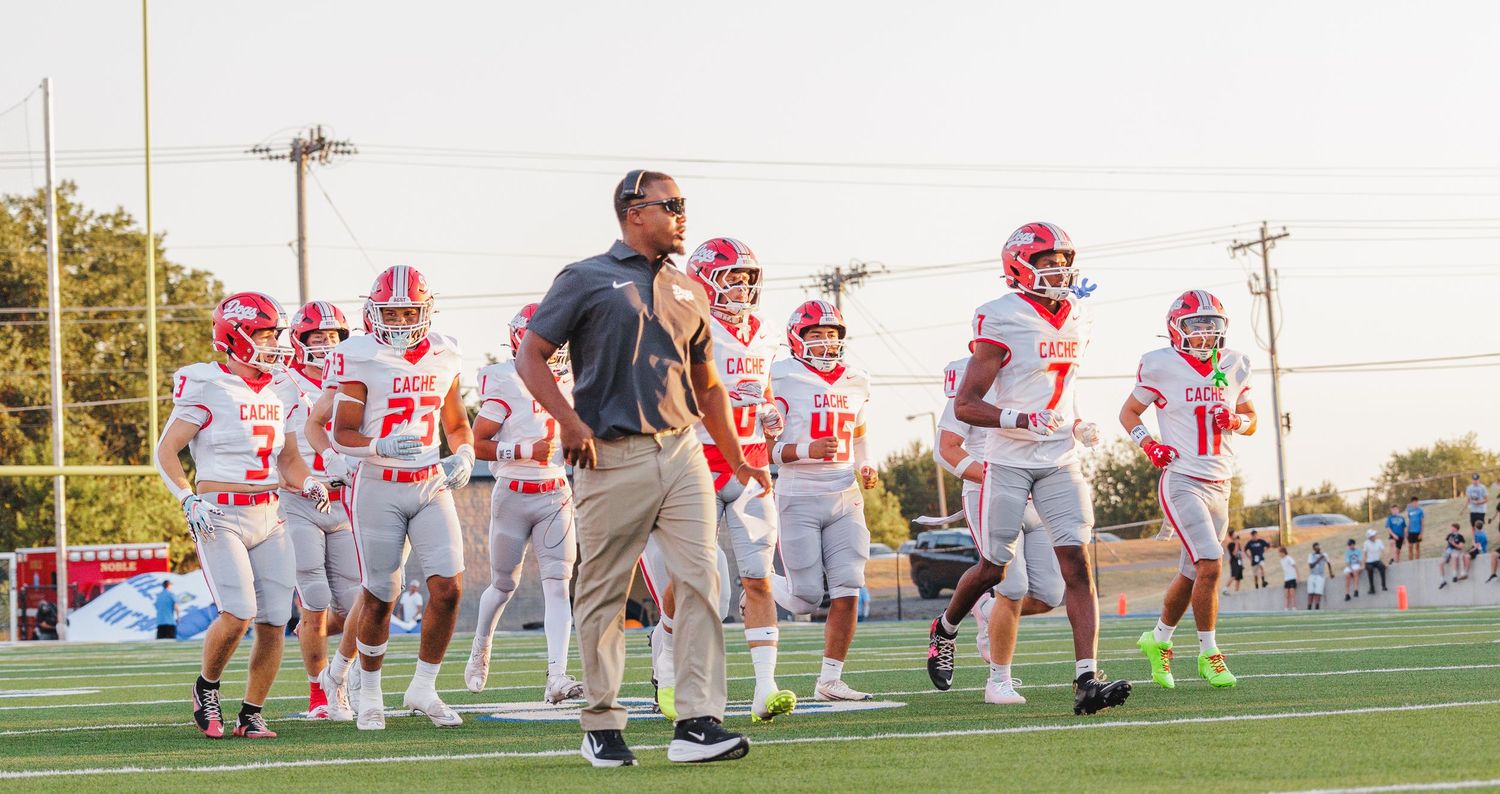 Football coach leads team onto field in red and white uniforms during an evening game under bright stadium lights.