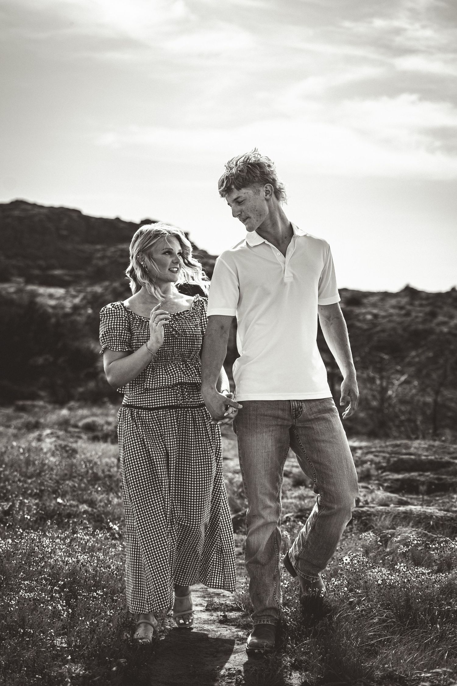 Black and white photo of a couple holding hands and walking outdoors on a rocky path under a cloudy sky.