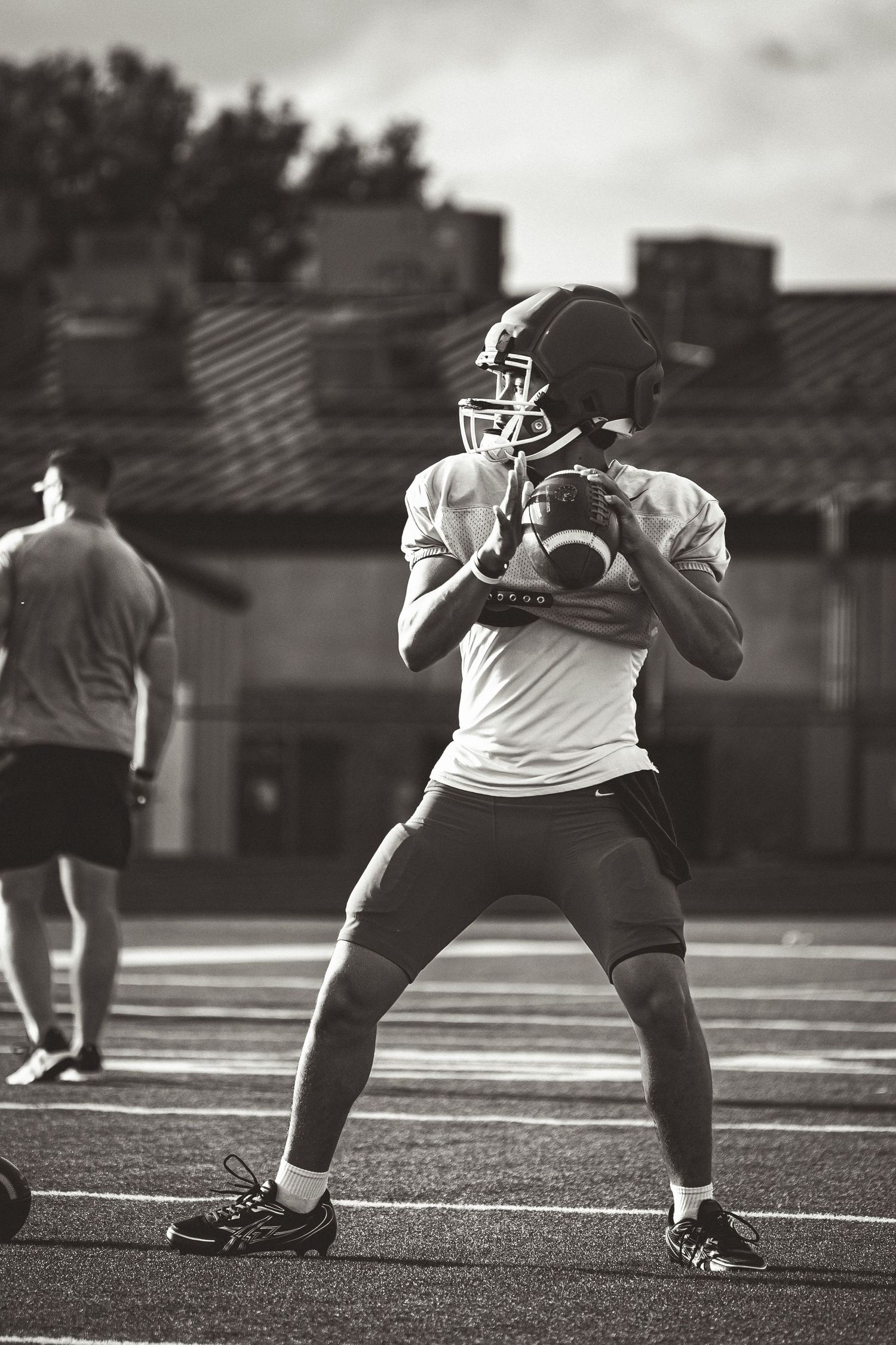 Black and white photo of a football player in helmet holding a ball in throwing stance on a practice field.