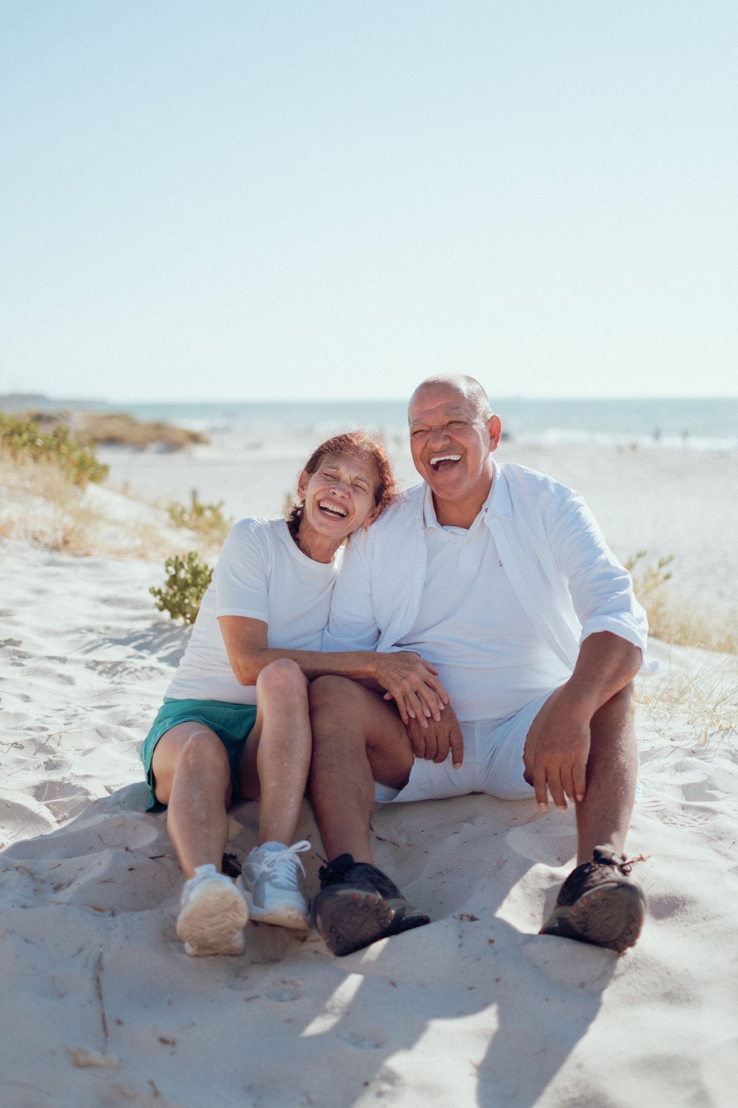 A couple sits together laughing on a sunny beach with white sand and blue ocean in the background.