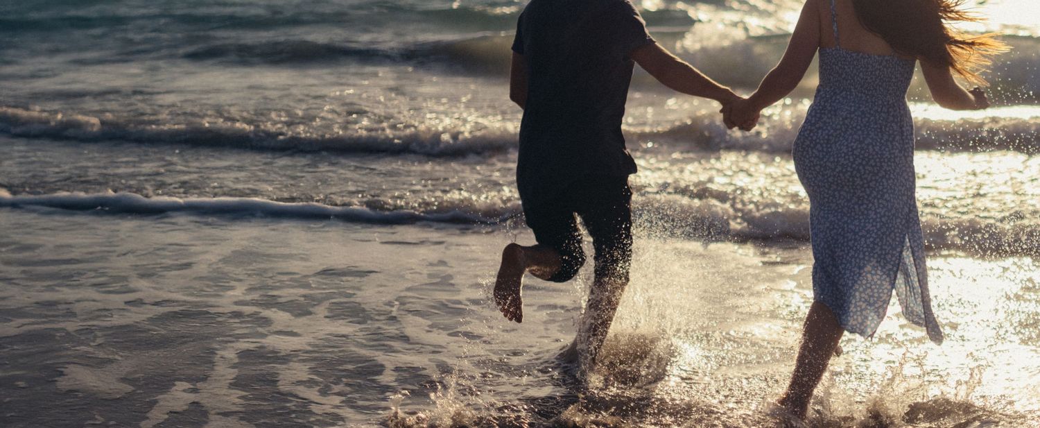 Couple holding hands while playing in ocean waves at sunset.