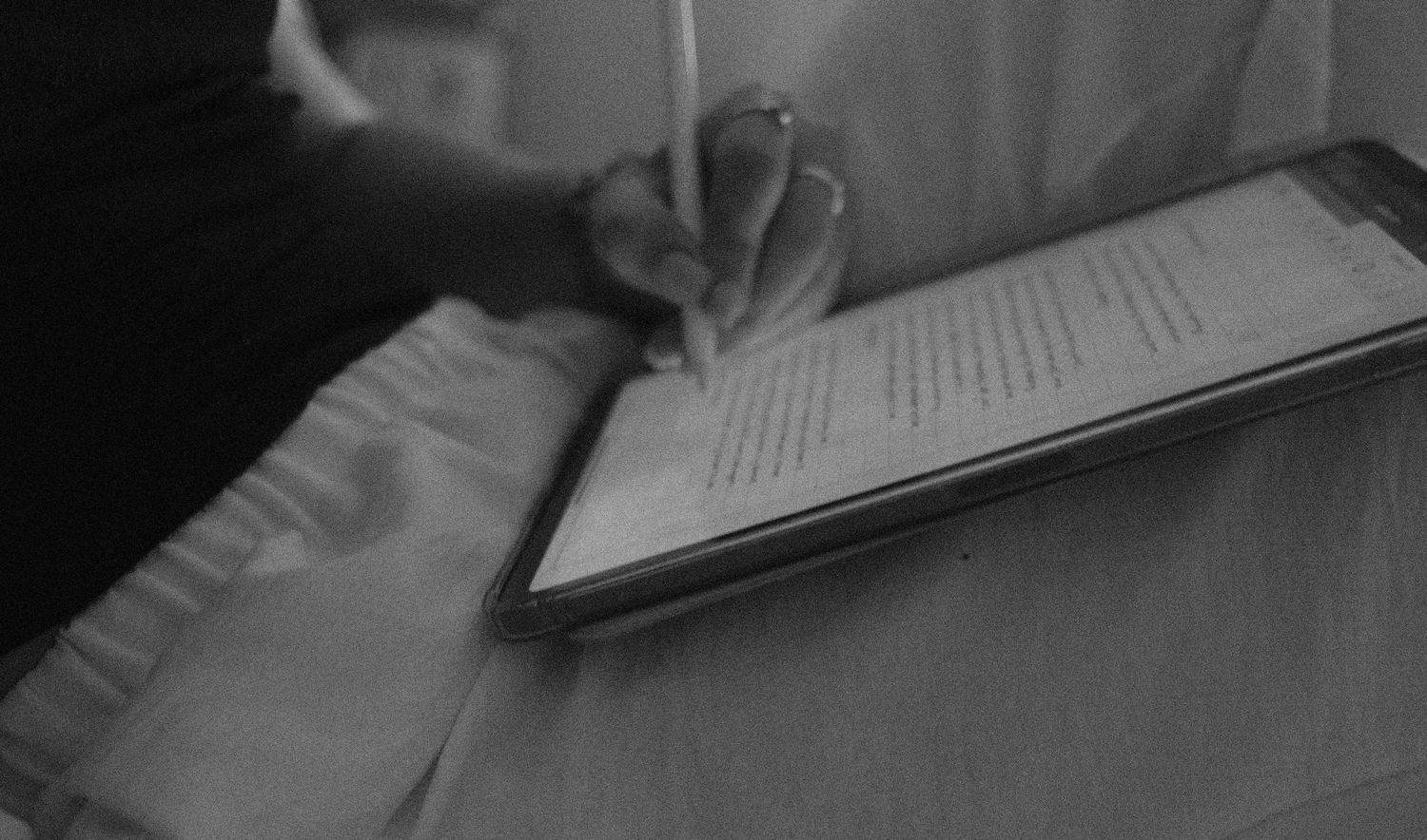 Black and white close-up of hands writing on paper in dim lighting.