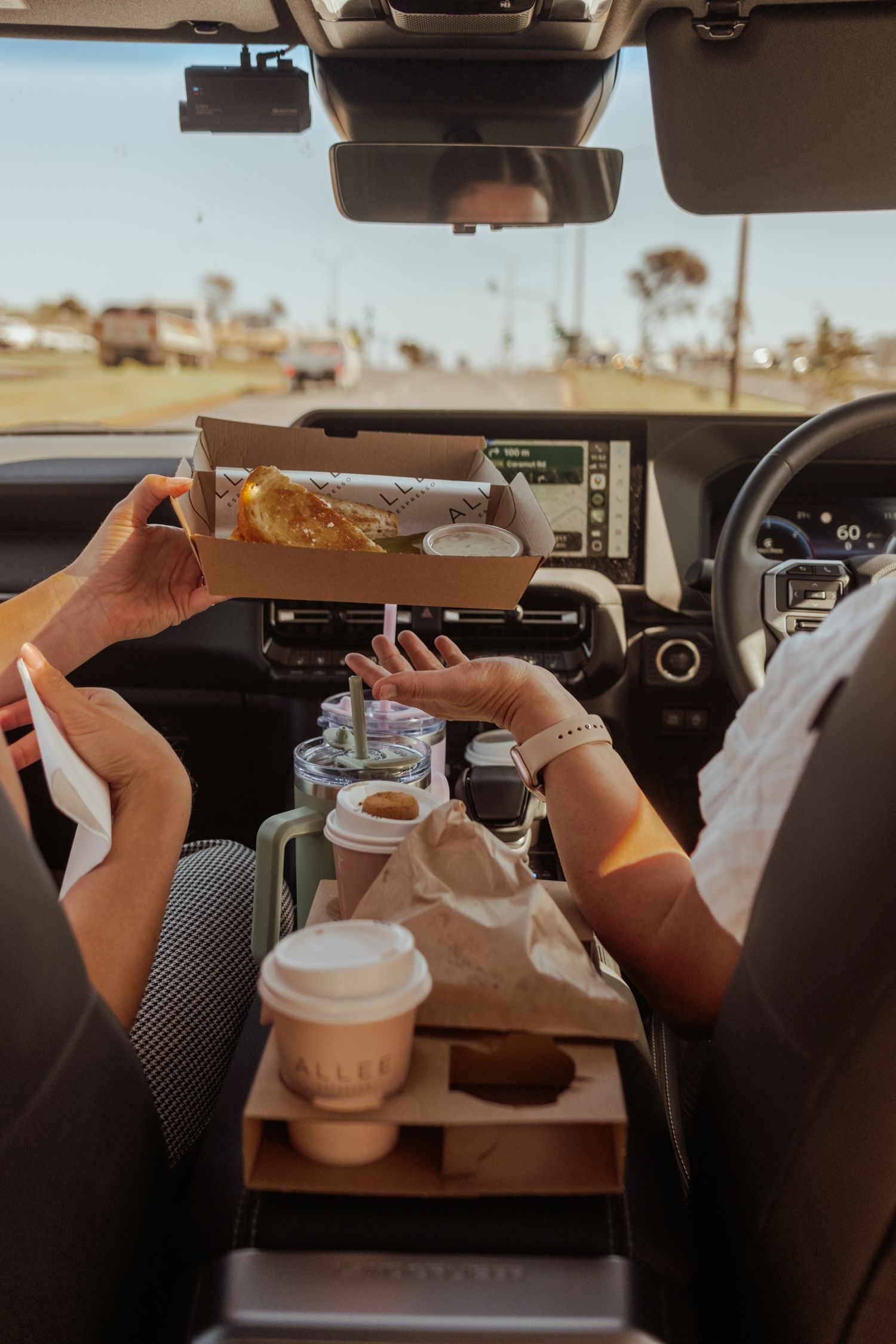 Car interior view showing hands holding takeout drinks and food on a tray.