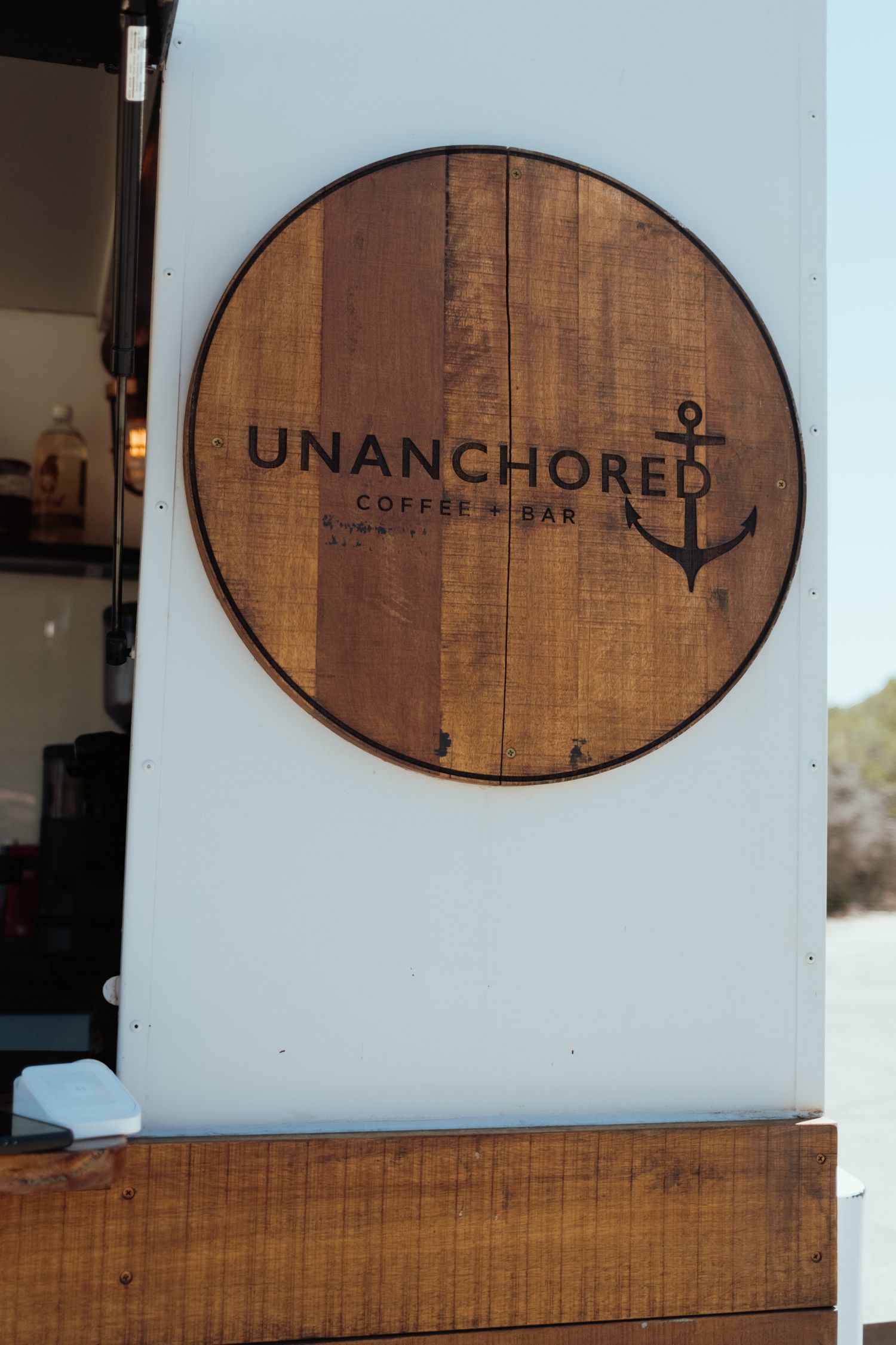 Wooden sign reading 'Unanchored Coffee & Bar' with anchor logo mounted on white wall.
