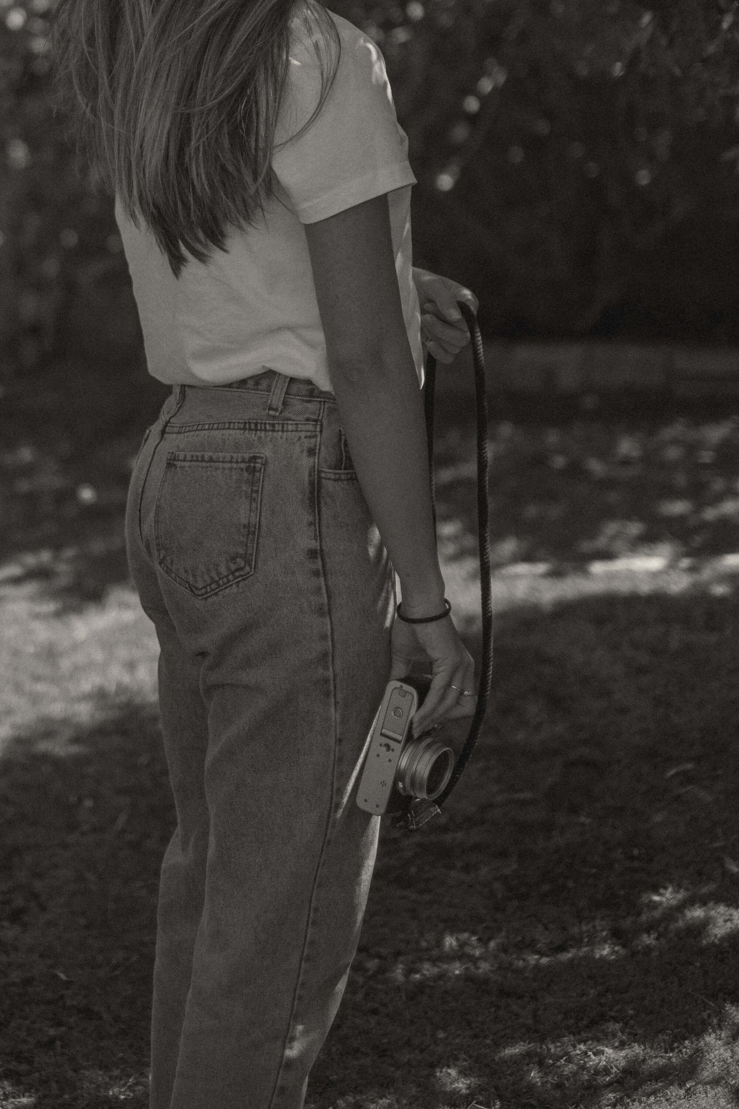 A person wearing jeans stands in a garden with dappled sunlight filtering through trees.