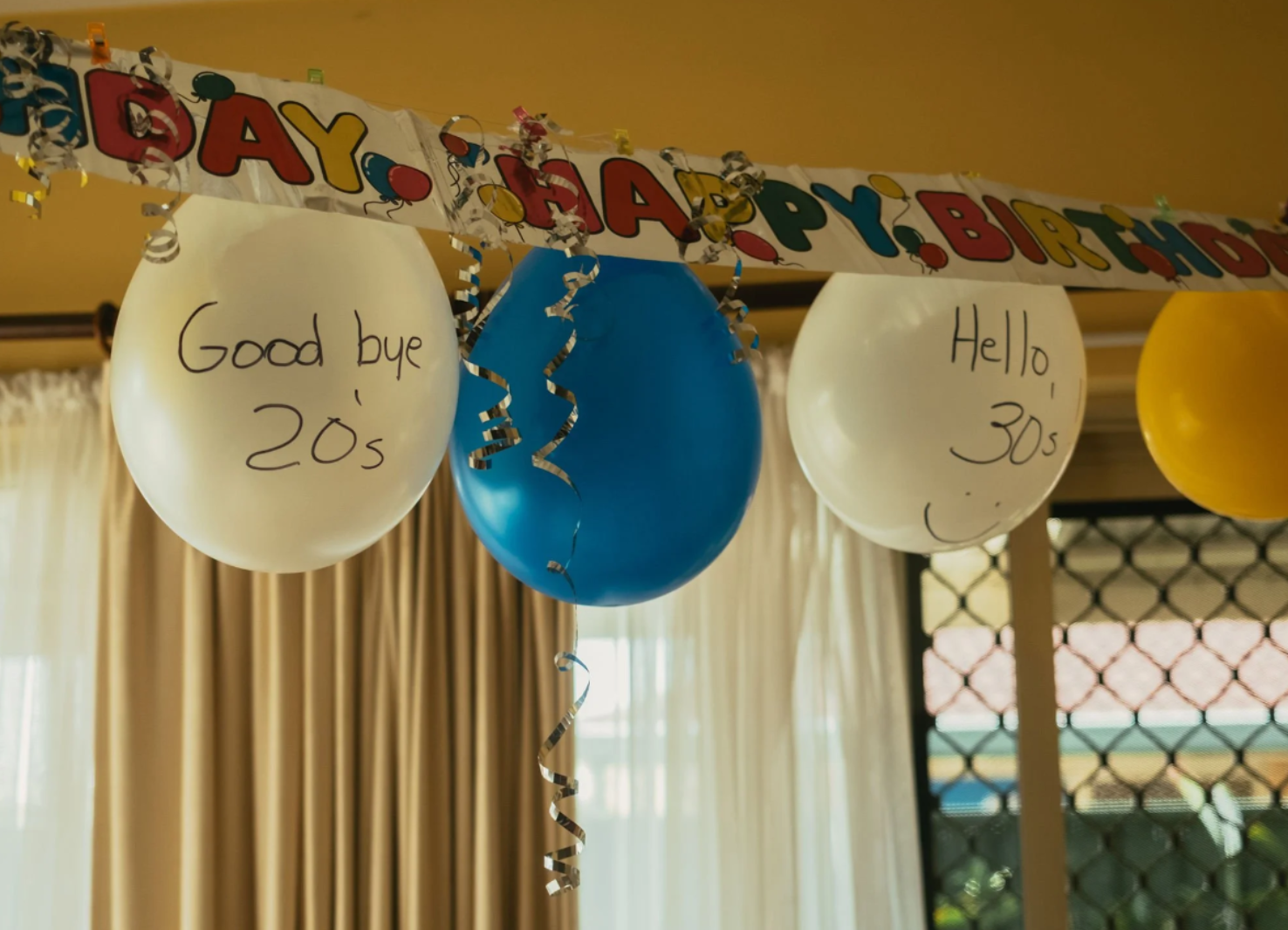 Colorful birthday balloons with messages written on them hanging against a yellow wall.