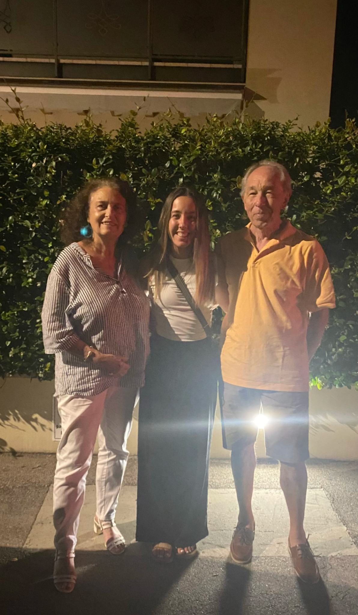 People posing for a photo outdoors at night with bushes in the background.