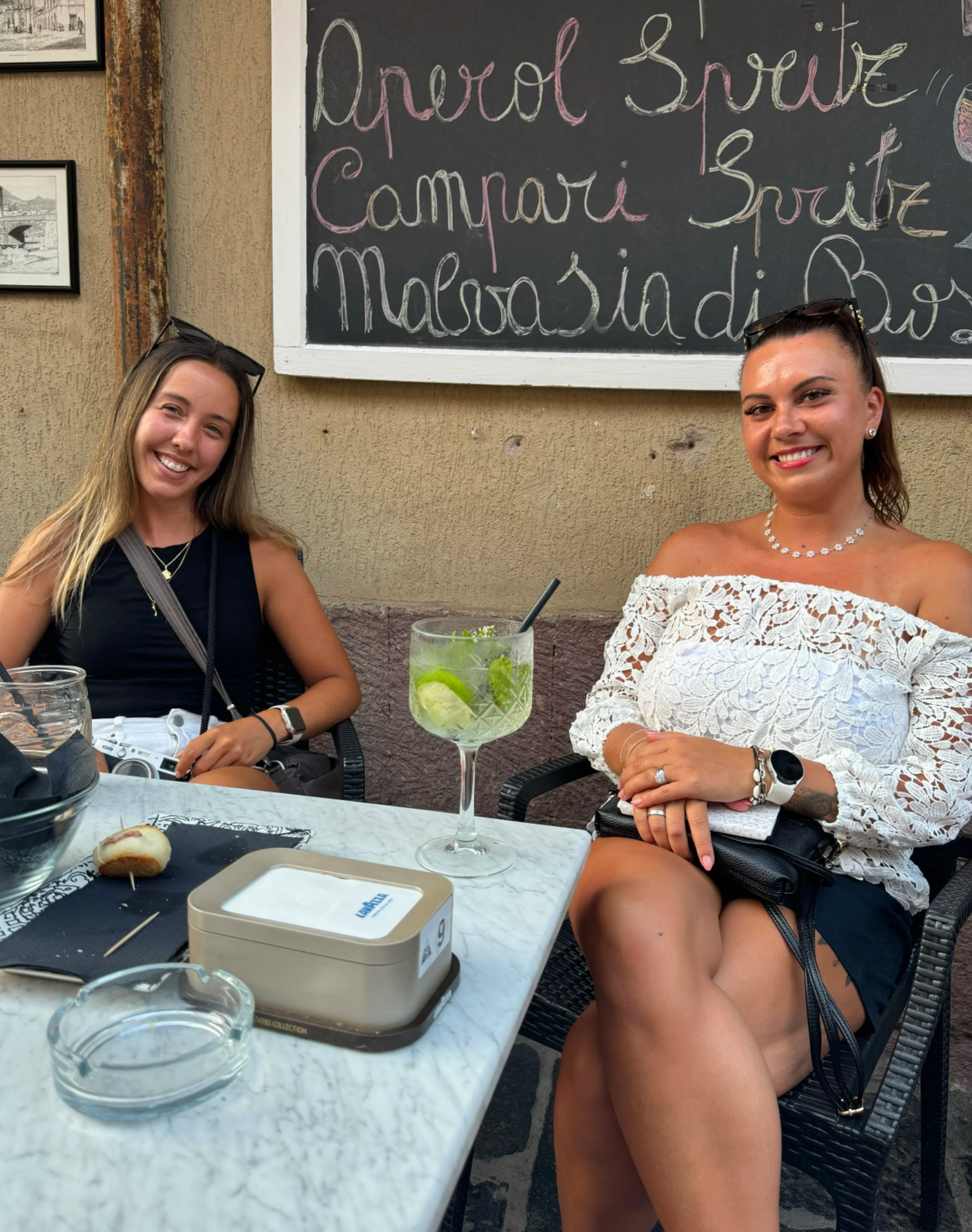 People sitting at an outdoor cafe table with drinks and a chalkboard menu visible in background.