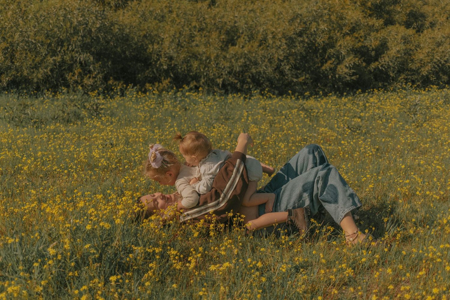 Mother lying in a field of yellow wildflowers on a sunny day with her children.