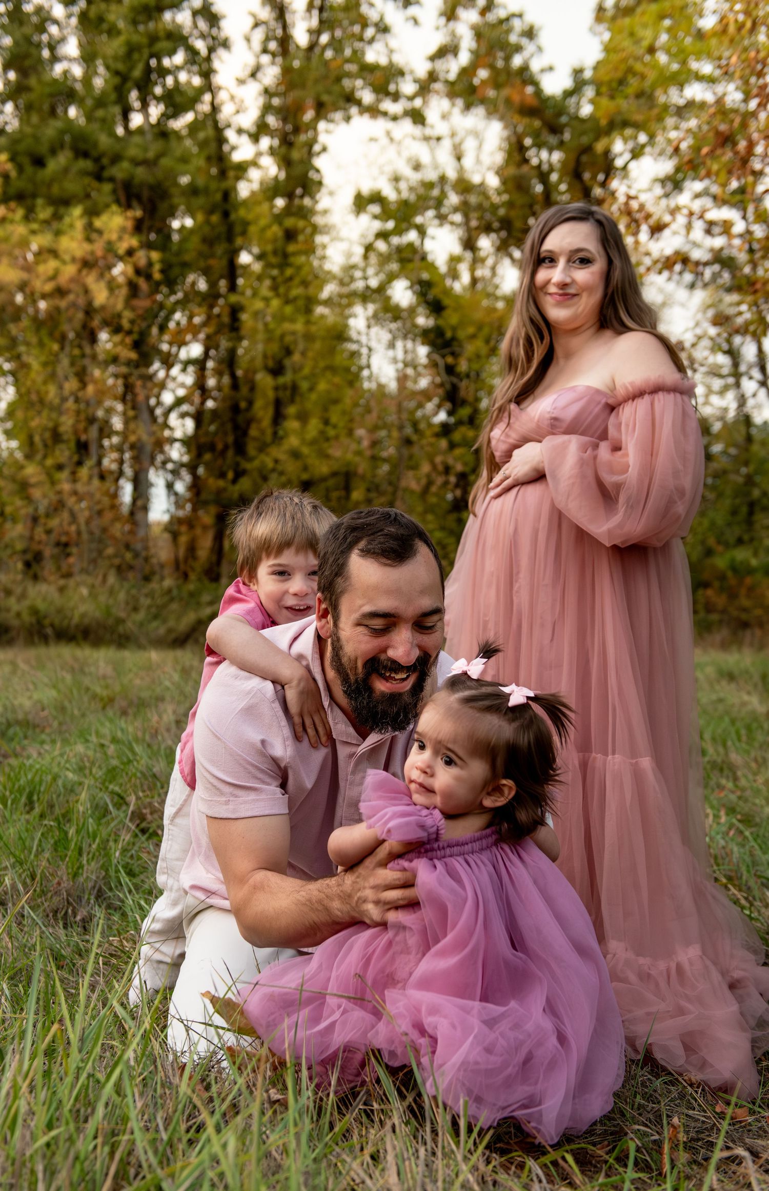 Family  in a field with trees in the background. Dad is playing with the kids on the ground while mom embraces her baby bump. Salem, OR.