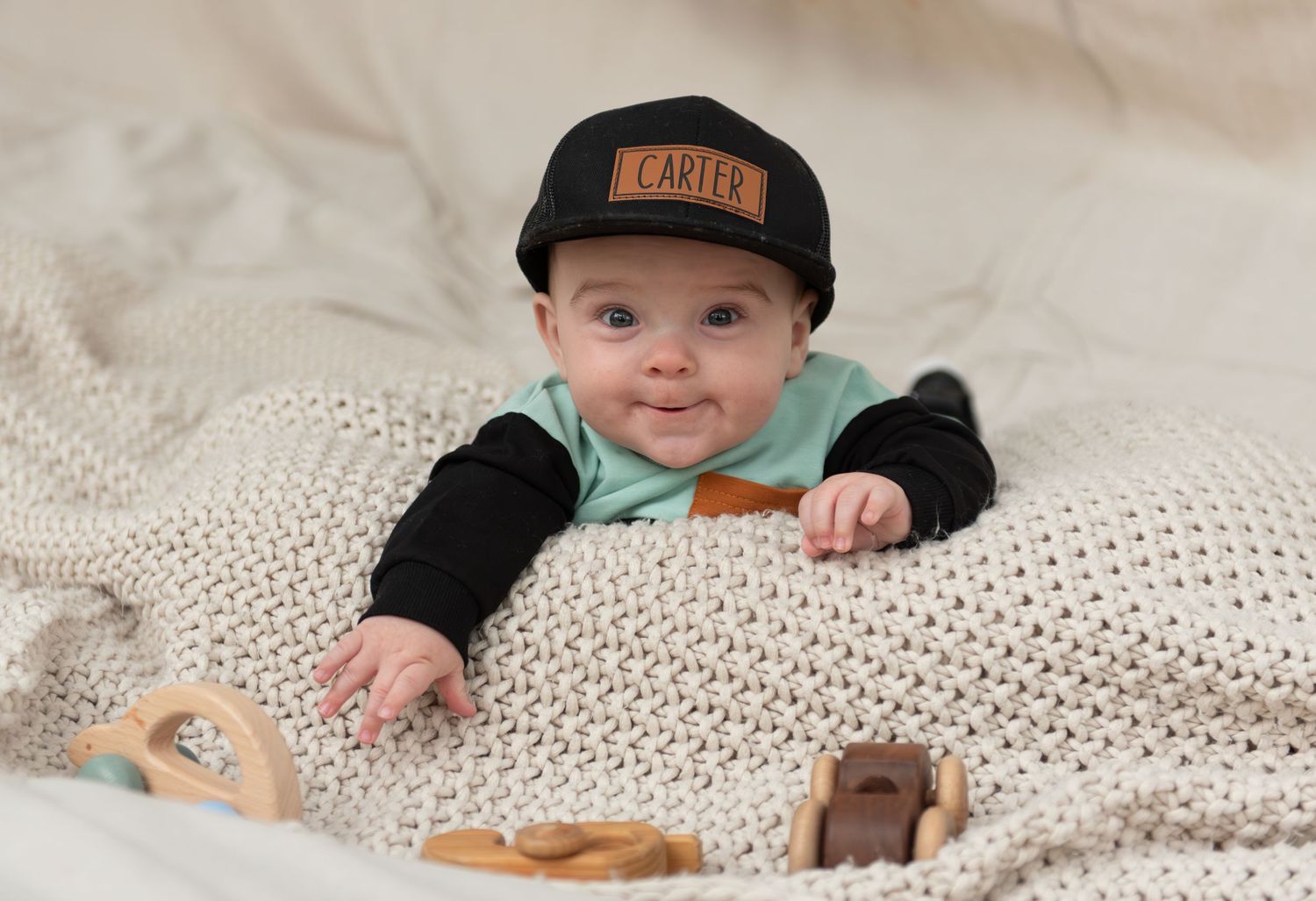 Six month milestone image with baby laying on a blanket, smiling and interacting with parents. Neutral backdrop with lettering. Salem, OR.