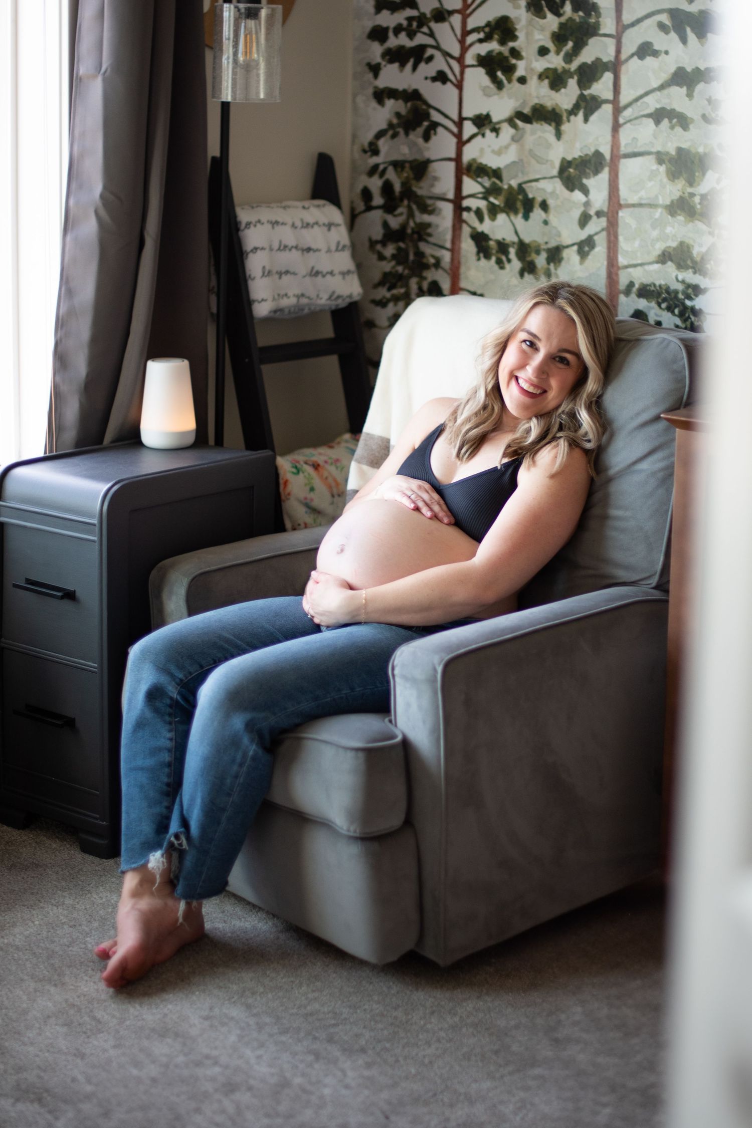 mother lounges comfortably on a gray armchair next to decorative vine plants in a cozy home setting during a maternity session. Located in S