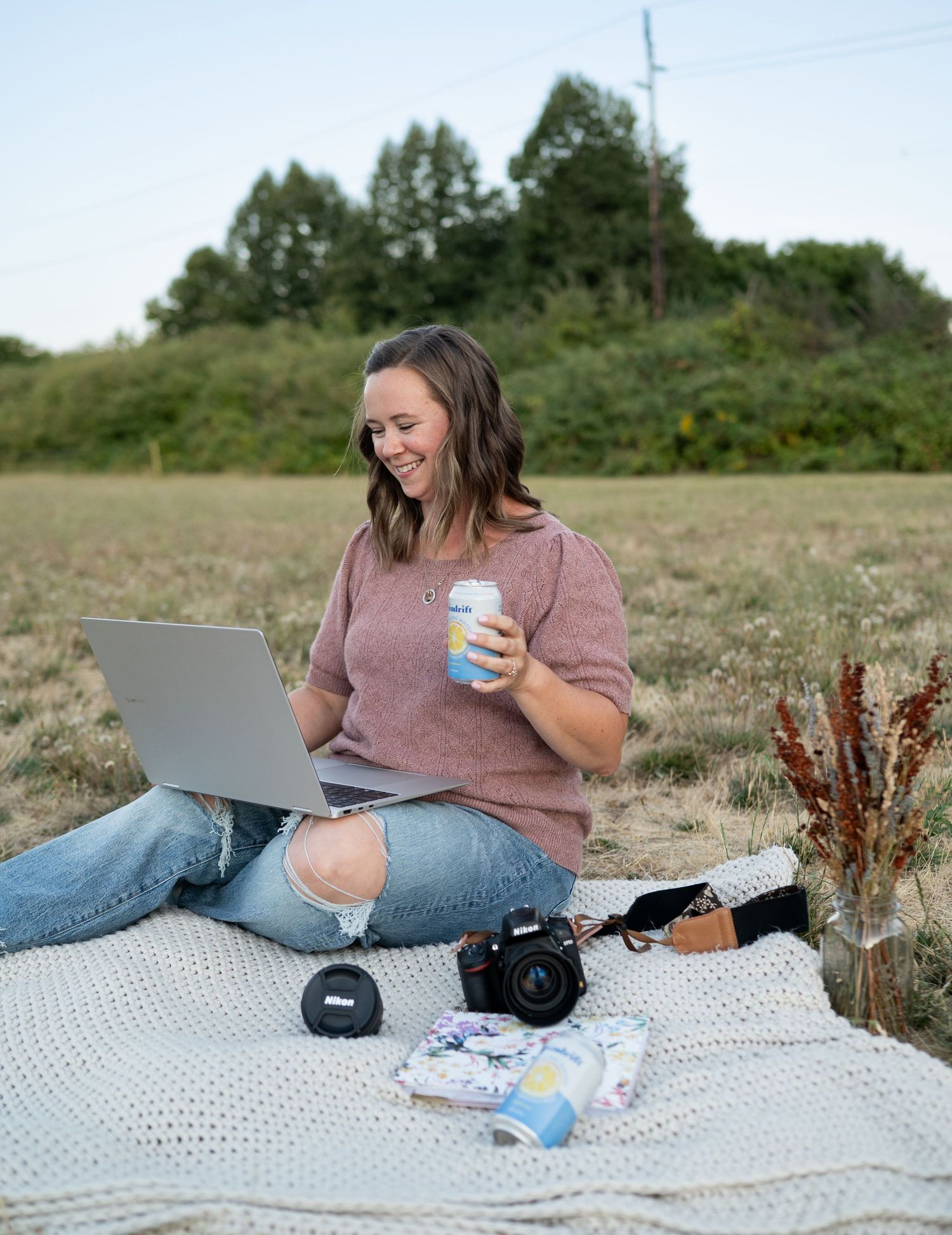 Someone enjoys working outdoors on a laptop while having coffee on a picnic blanket with camera and dried flowers nearby.