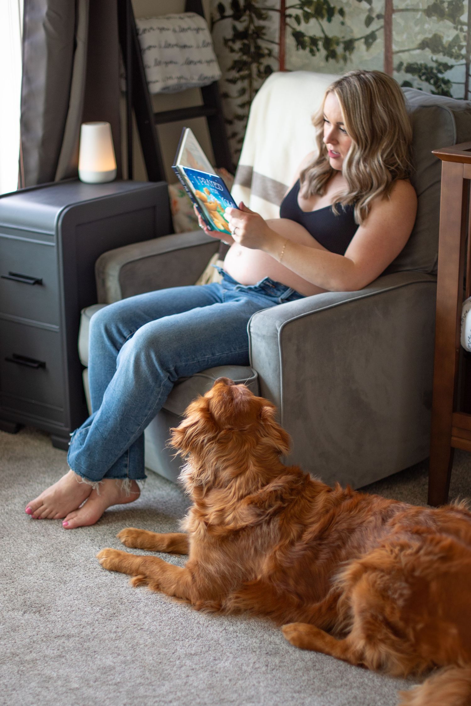 A golden retriever dog lies on carpet next to a mother sitting in a gray armchair reading a book in her baby's nursery. Located in Salem, OR