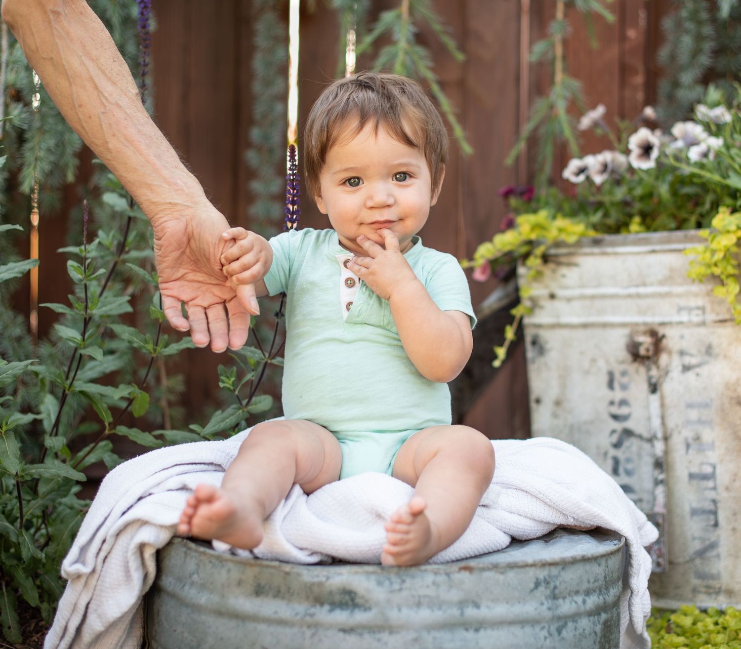 A small child in mint green outfit sits on a galvanized metal tub in a garden setting while holding an adult's hand.