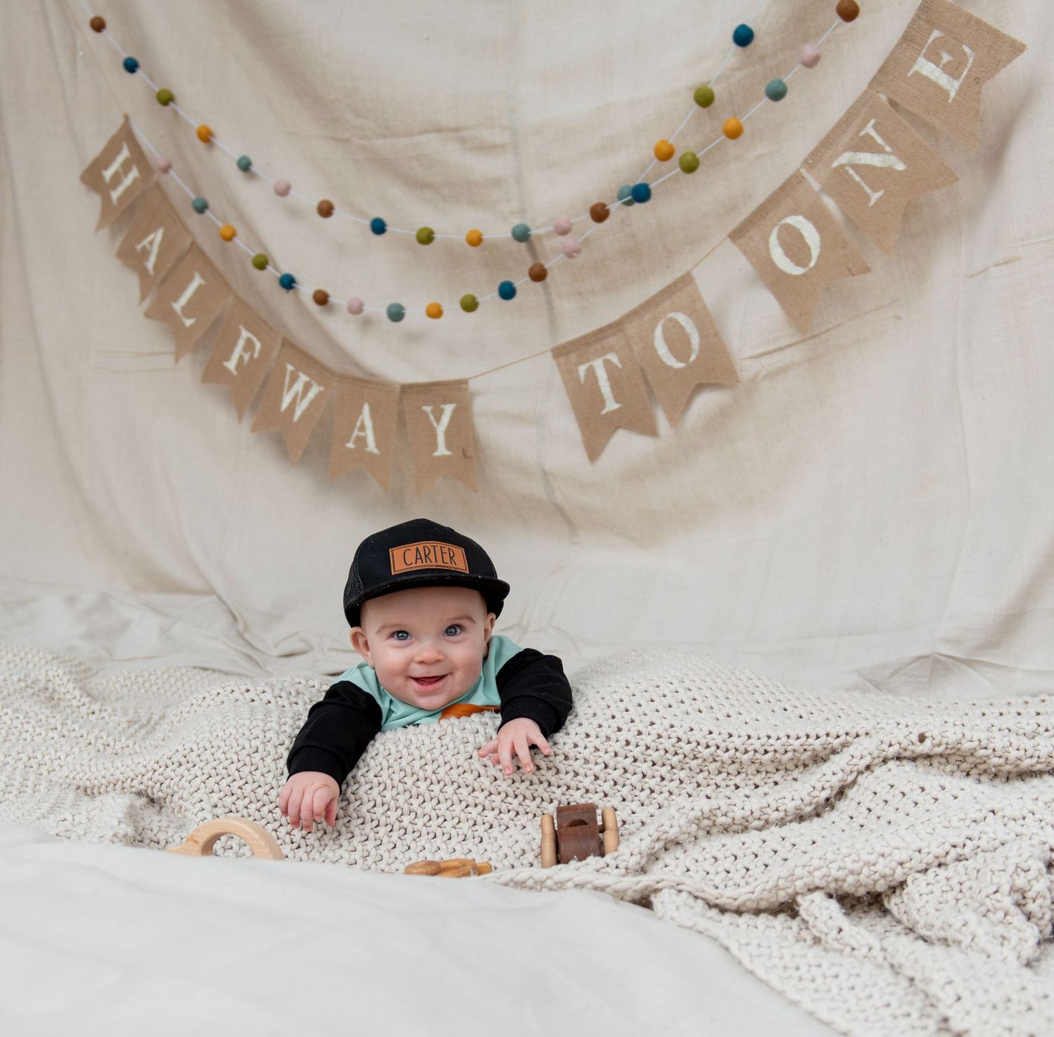 Six month milestone image with baby laying on a blanket, smiling and interacting with parents. Neutral backdrop with lettering. Salem, OR.