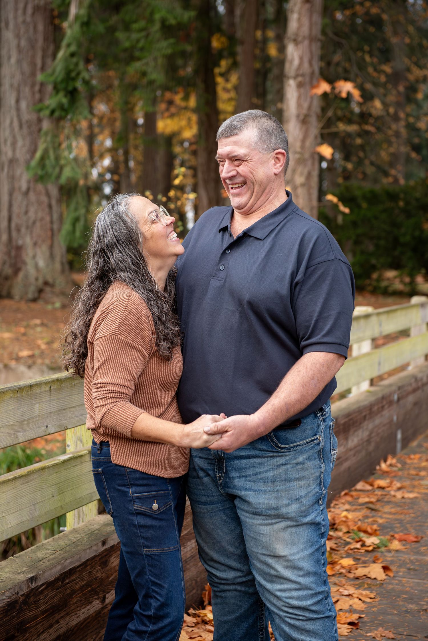 A couple shares joyful laughter while leaning against a wooden bridge surrounded by autumn leaves in a park setting. located in Stayton, OR
