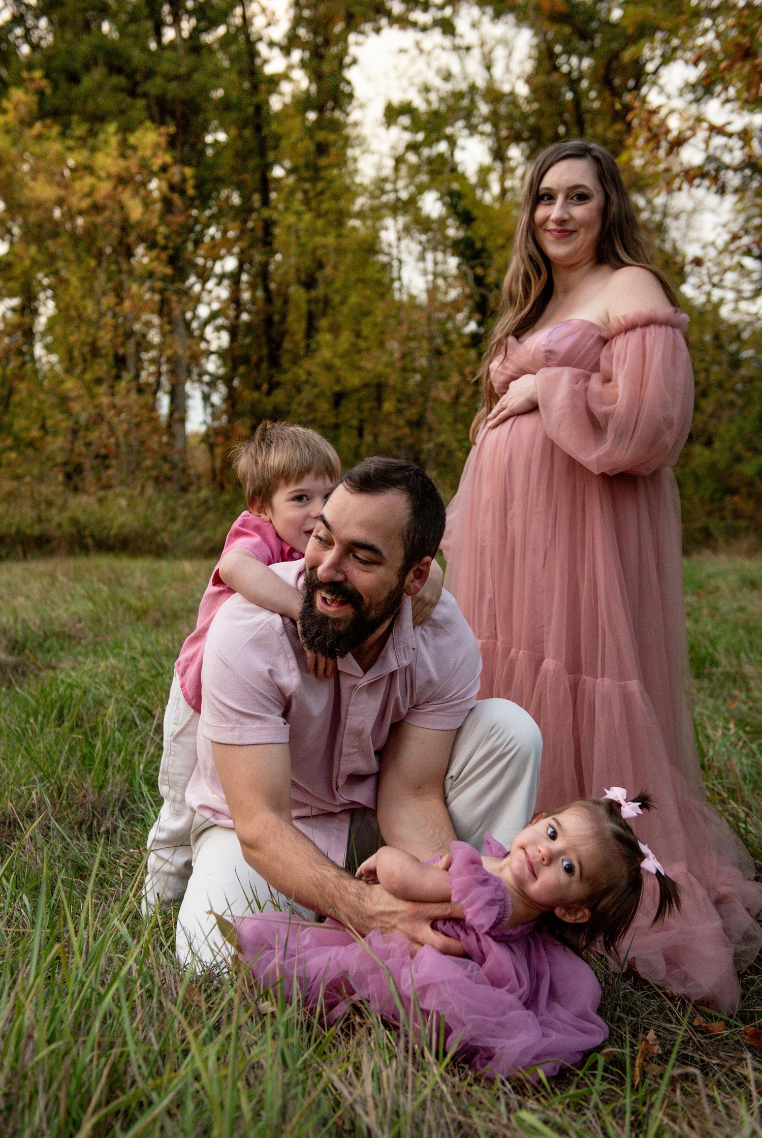 Family  in a field with trees in the background. Dad is playing with the kids on the ground while mom embraces her baby bump. Salem, OR