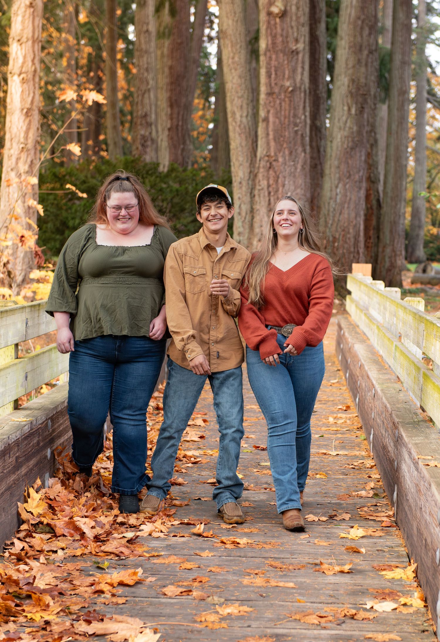 Three siblings walk along a wooden bridge covered in fallen autumn leaves through a forest of tall trees. Stayton, OR
