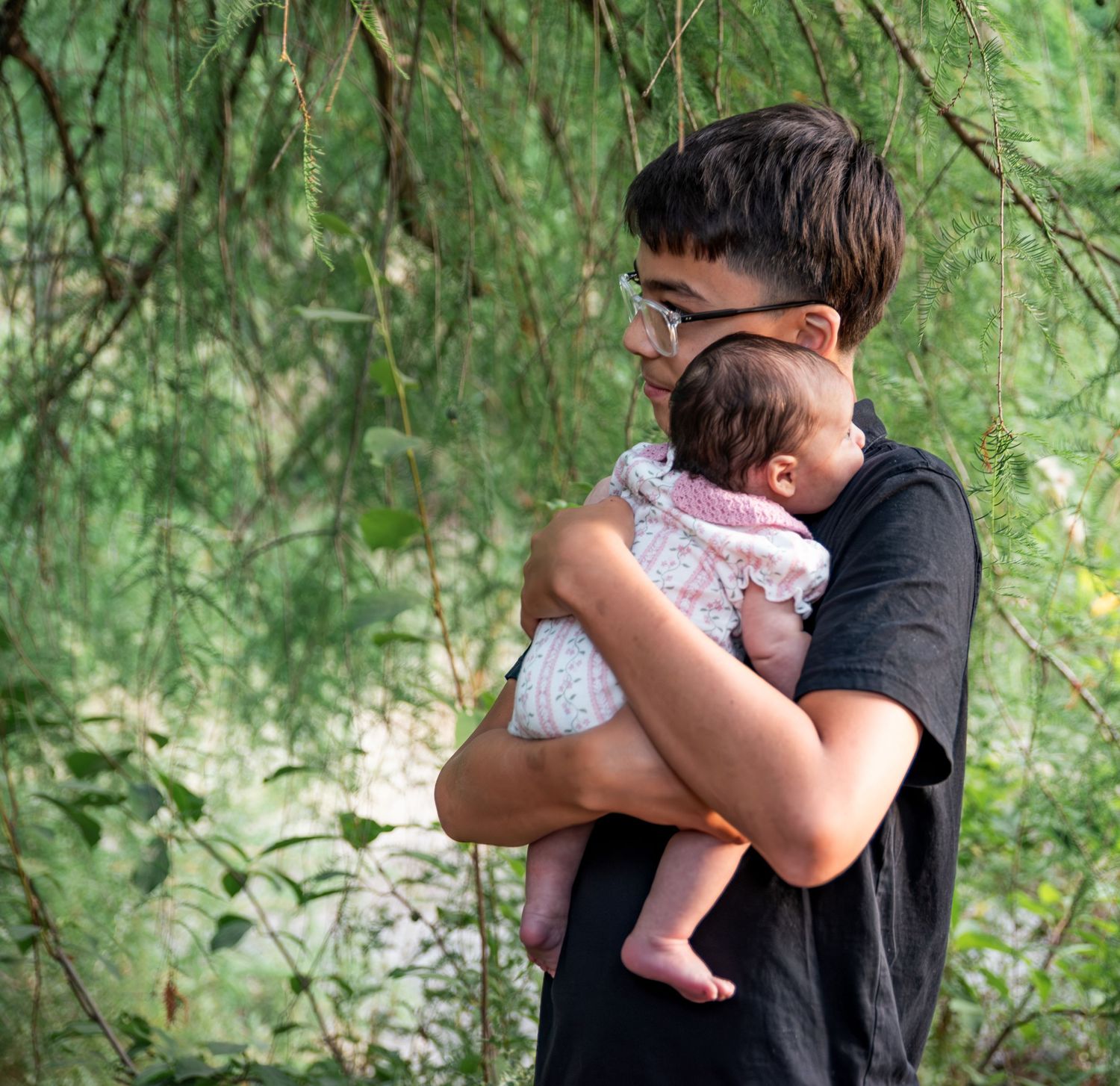 Older brother embracing infant sister under a willow tree located in Salem, OR.